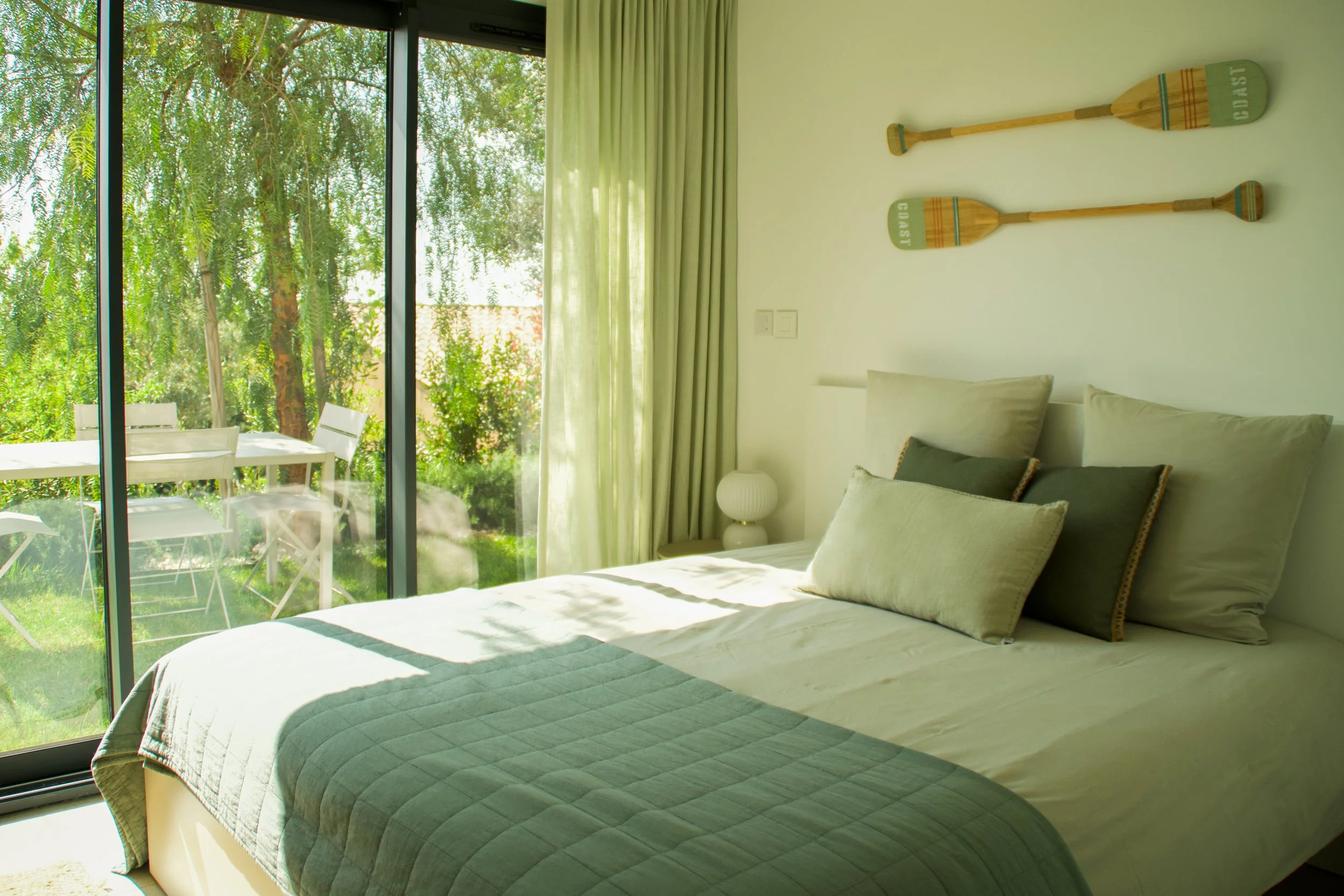 Photographie AirBnB Immobilier. Chambre lumineuse avec lit fait, vue sur un jardin vert à travers une grande porte-fenêtre. Décorations murales en forme de pagaies en bois.