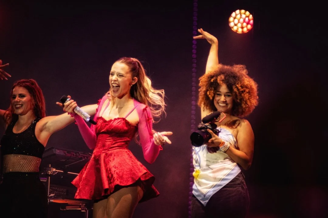 Photographe concert - Fête avec des femmes qui chantent et prennent des photos sur scène, ambiance dynamique et colorée.