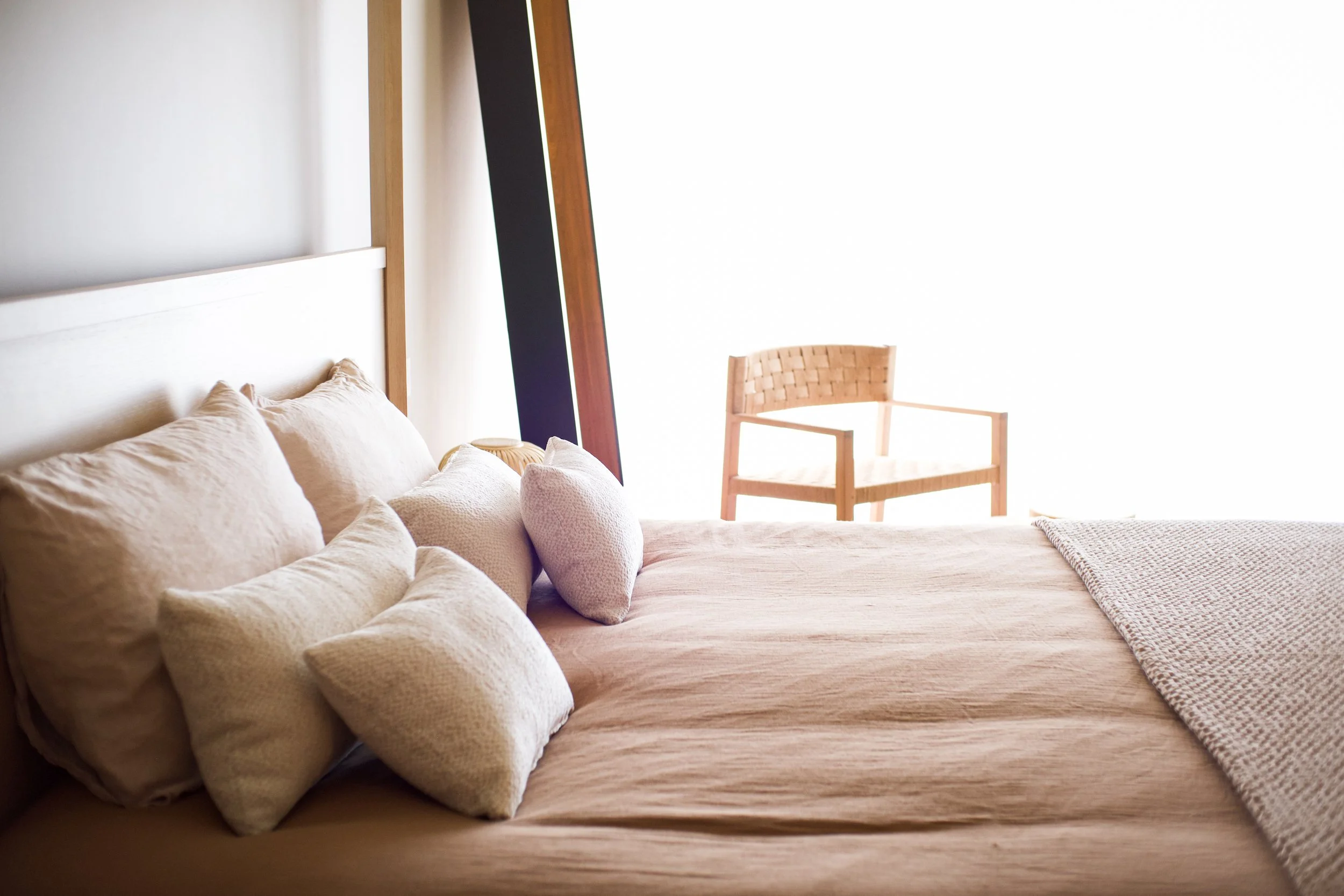 Photographie AirBnB Immobilier. Lit avec plusieurs coussins en tissus beige clair, face à un mur blanc. Une chaise en bois avec assise en tressage est visible à l'arrière, devant une grande fenêtre lumineuse.