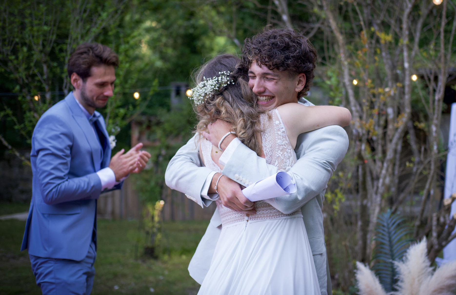 Photographe mariage - Deux personnes, un homme et une femme, se font un câlin lors d'une célébration en plein air, probablement un mariage, avec un homme en costume gris en arrière-plan applaudi.