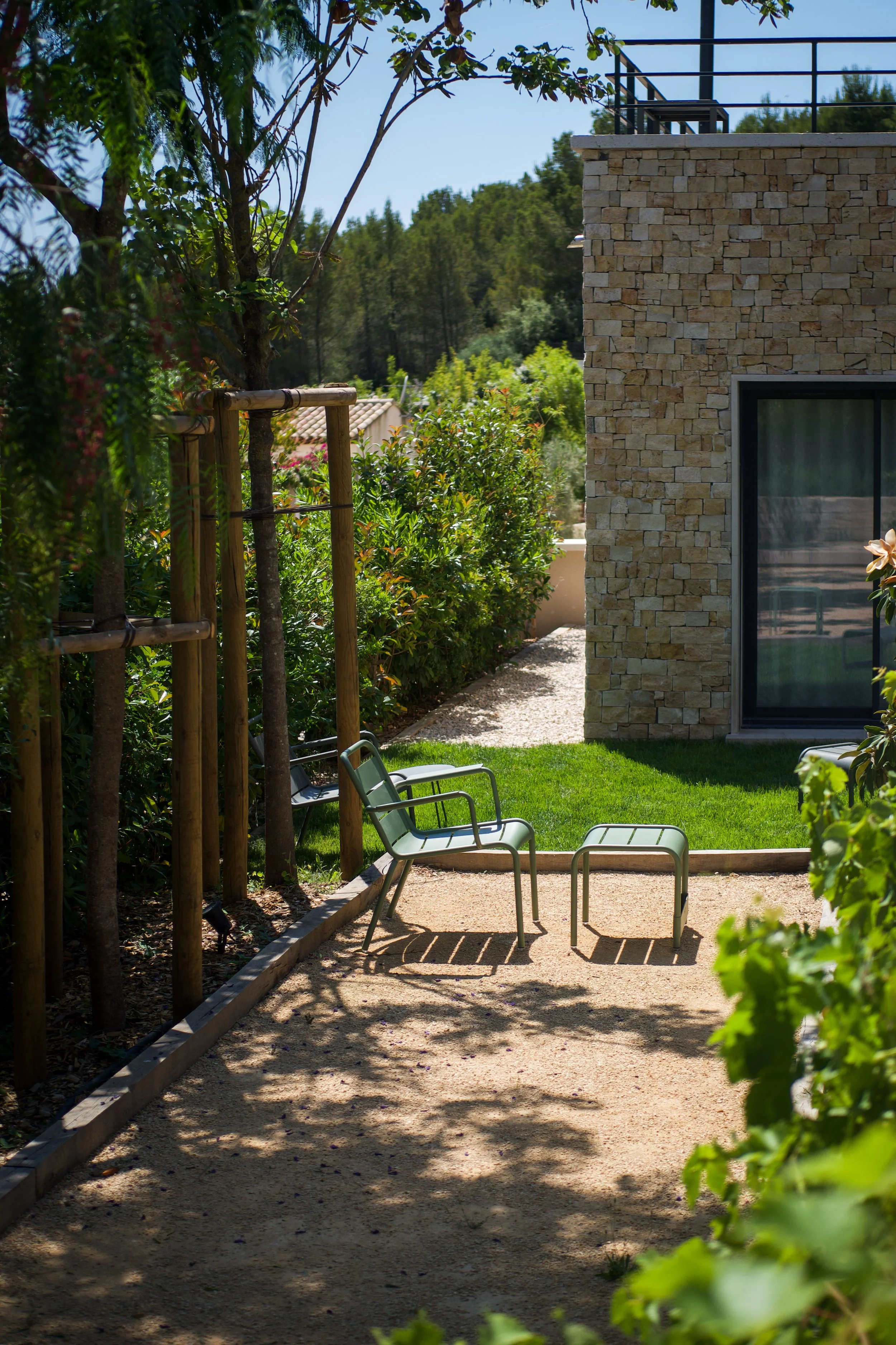 Photographie AirBnB Immobilier. Un coin de jardin avec deux chaises en métal vert clair et un petit banc en métal, entouré de végétation et à côté d'une maison avec un mur en pierre, sous un ciel ensoleillé.