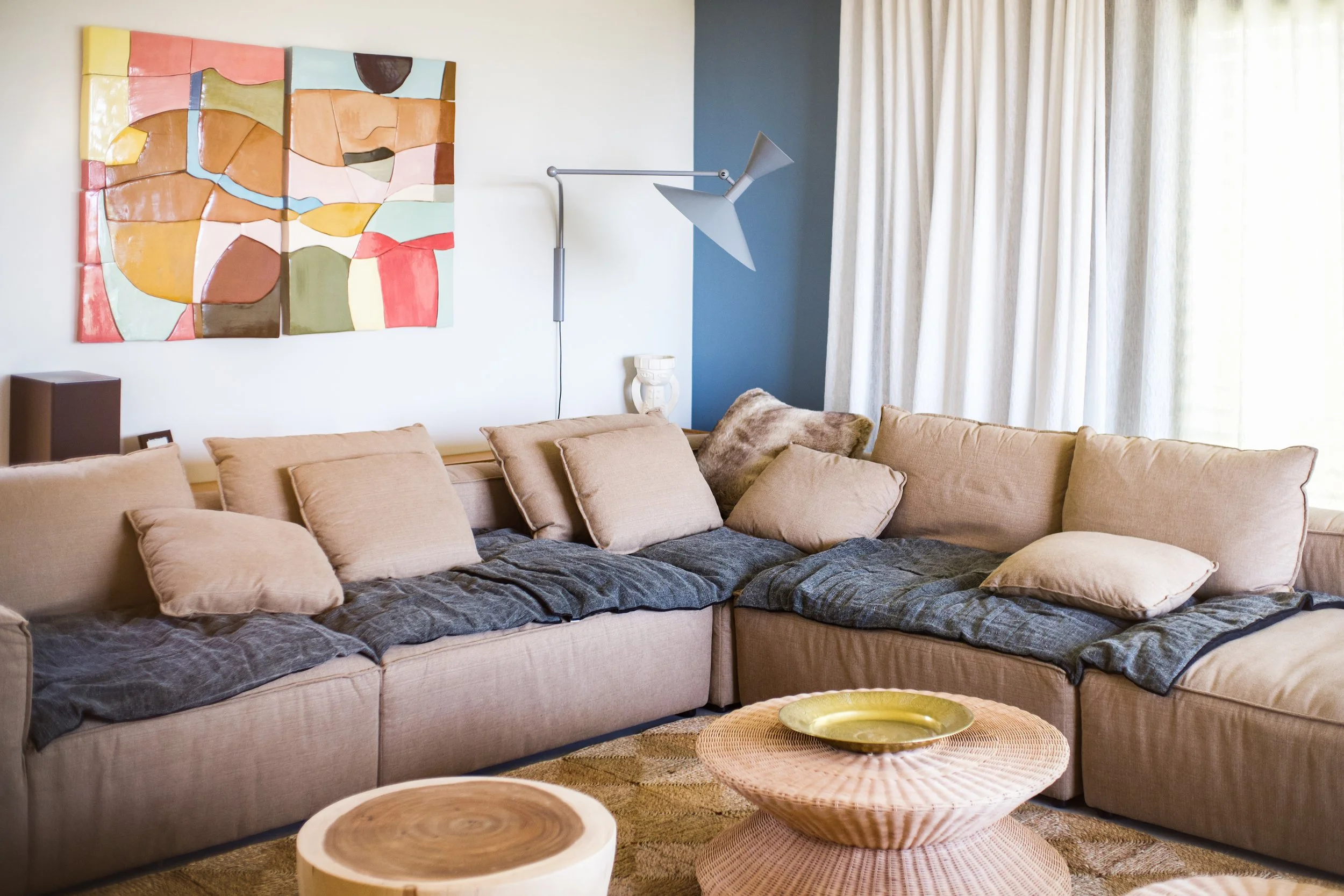 Photographie AirBnB Immobilier. Salon avec canapé beige, coussins, peinture abstraite, lampe moderne, rideaux blancs, table en rotin, décoration d'intérieur