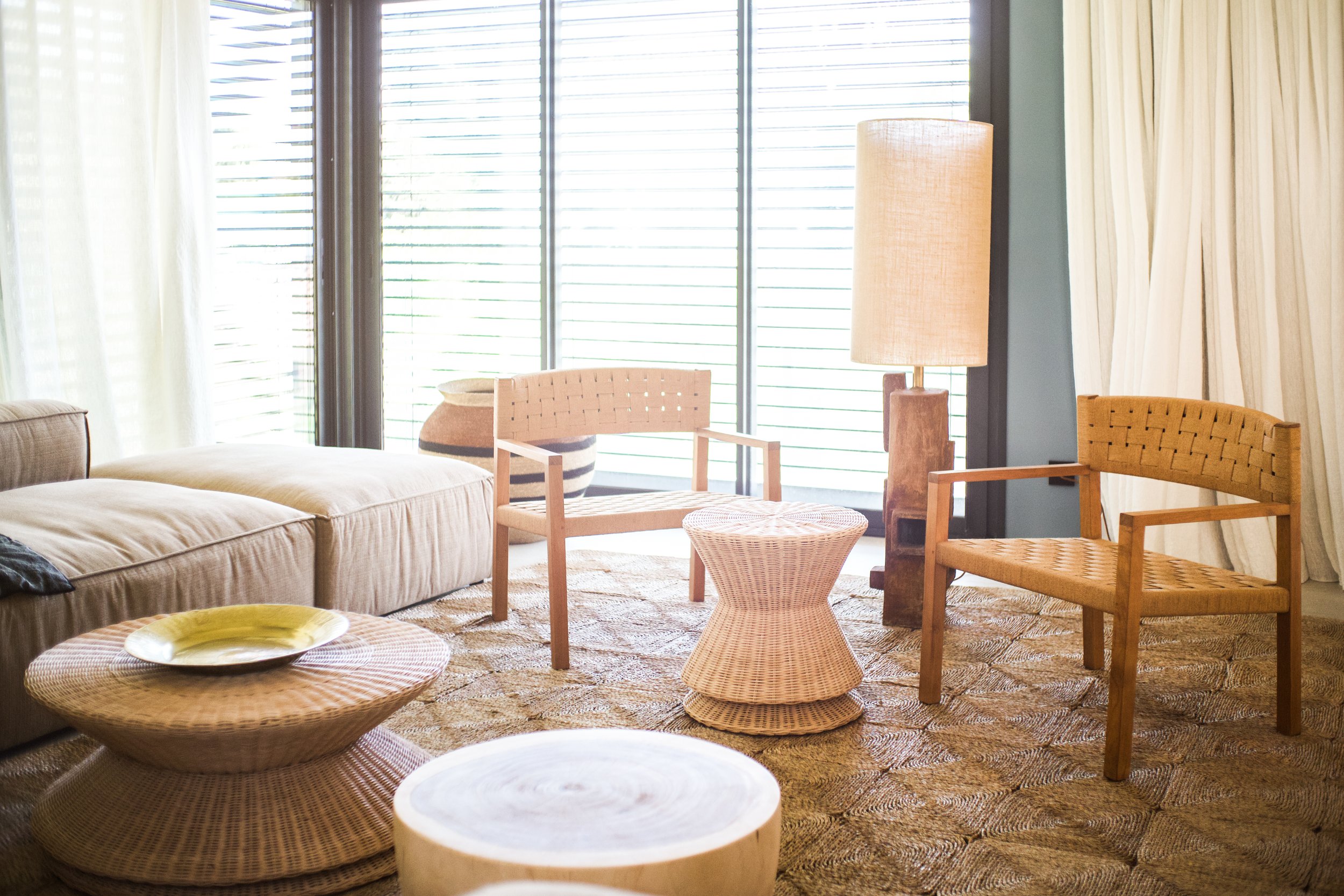Photographie AirBnB Immobilier. Salon avec deux chaises en bois, un canapé beige, un lampadaire, une table en rotin, une grande fenêtre avec volets, et une tapis en fibre naturelle.