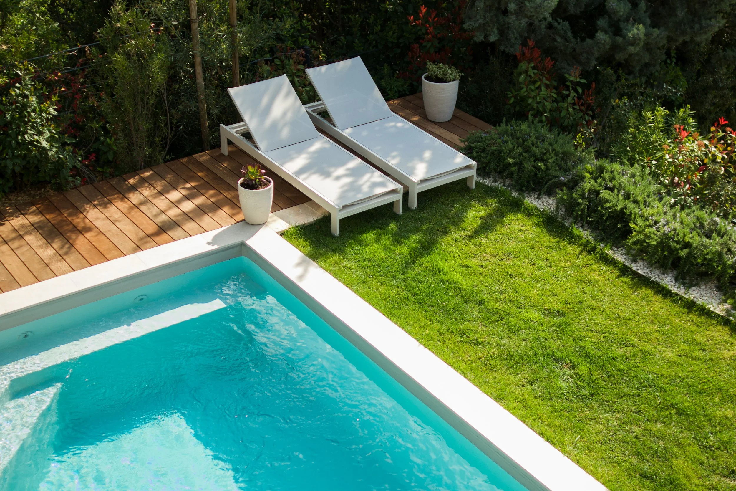 Photographie AirBnB Immobilier. Piscine avec deux chaises longues blanches, entourée de pelouse verte, avec des plantes en pot et un jardin derrière.