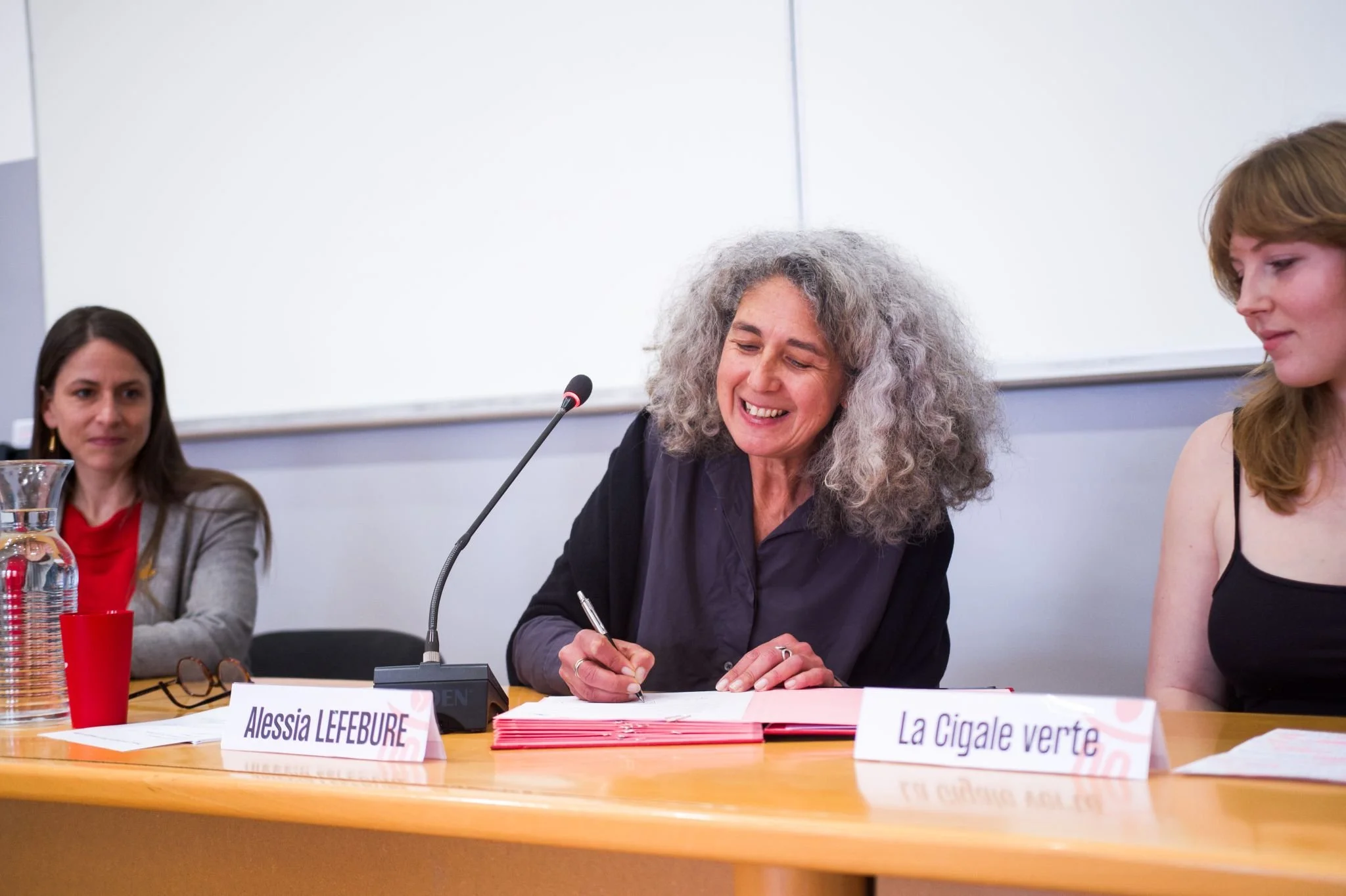 Photographe conférence - Une femme avec de longs cheveux bouclés gris, souriante, assise à une table, en train de signer un document. 