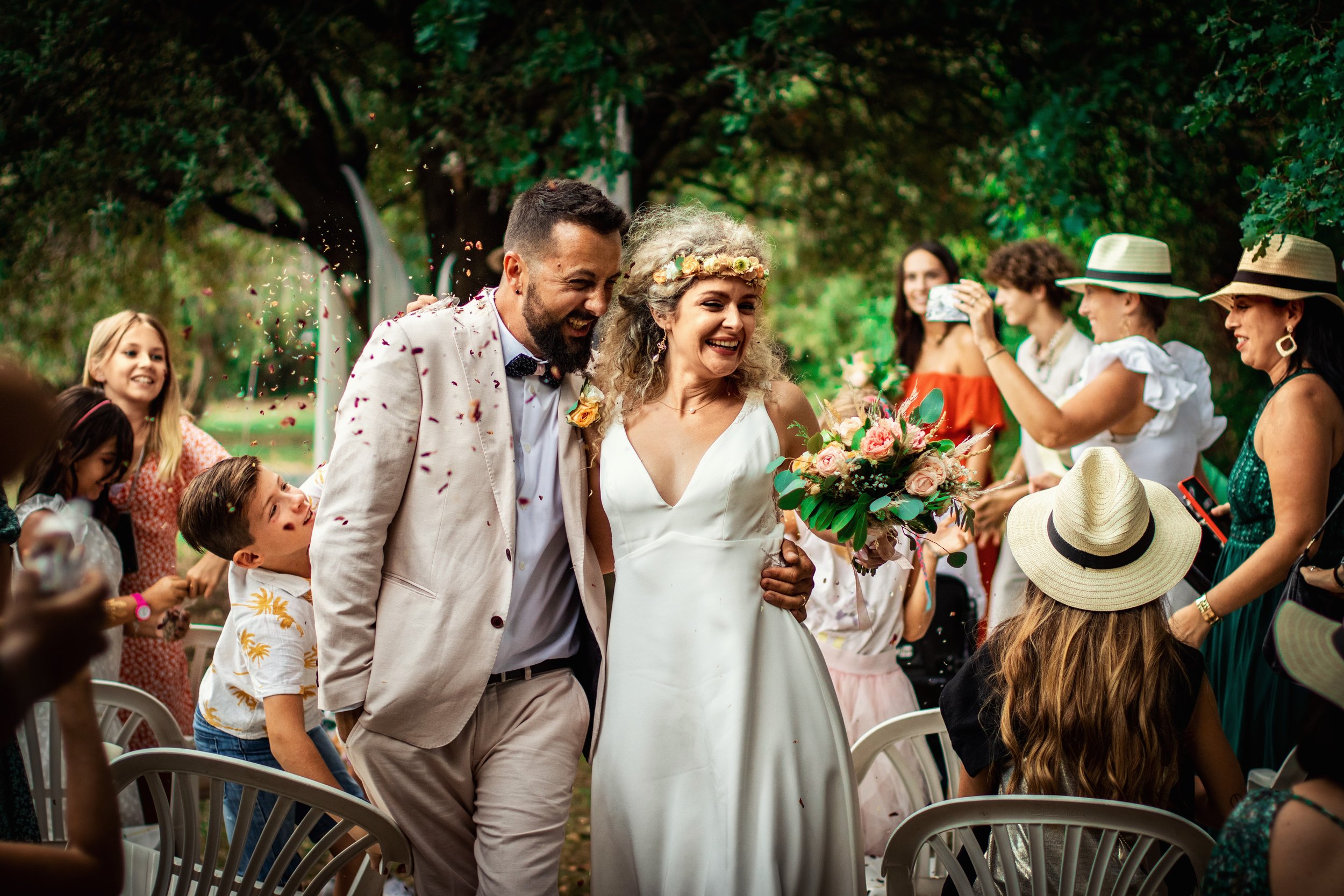 Photographe mariage - Un couple de mariés souriant, entouré d'invités lors d'un mariage en plein air, avec des invités prenant des photos et lançant des confettis.