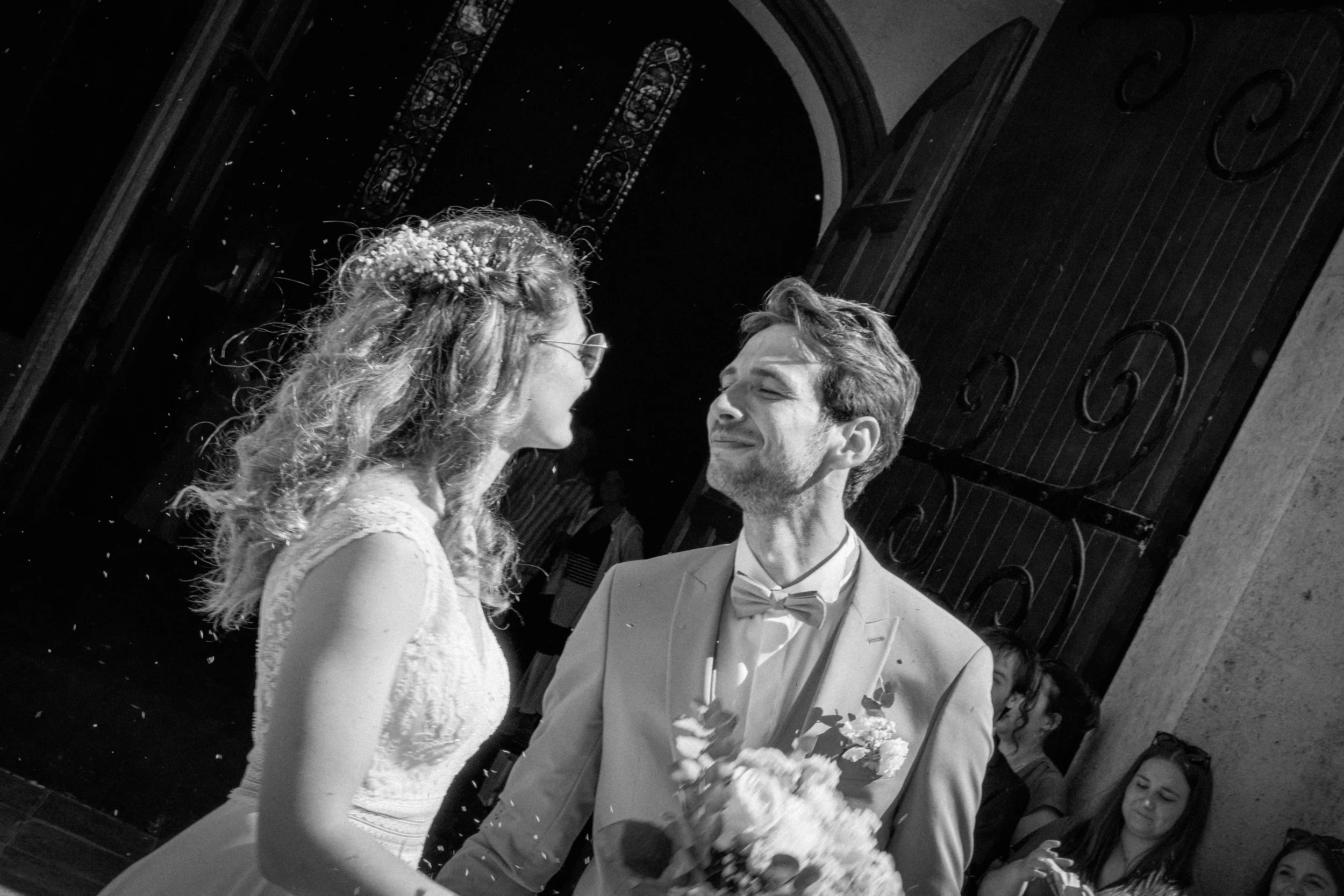 Photographe mariage - Un couple de jeunes mariés lors de leur mariage, en noir et blanc, souriant et regardant dans les yeux l'un de l'autre, à l'entrée d'une cérémonie.