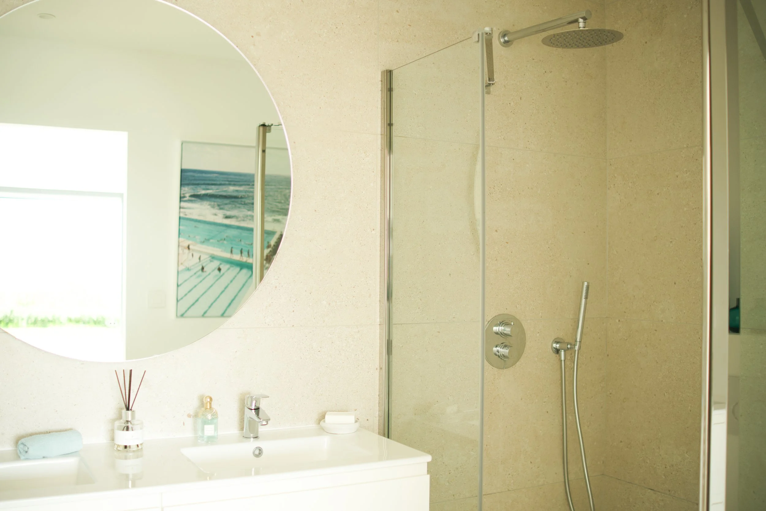 Photographie AirBnB Immobilier. Salle de bain moderne avec douche à l'italienne, miroir rond, lavabo, et décoration minimaliste.