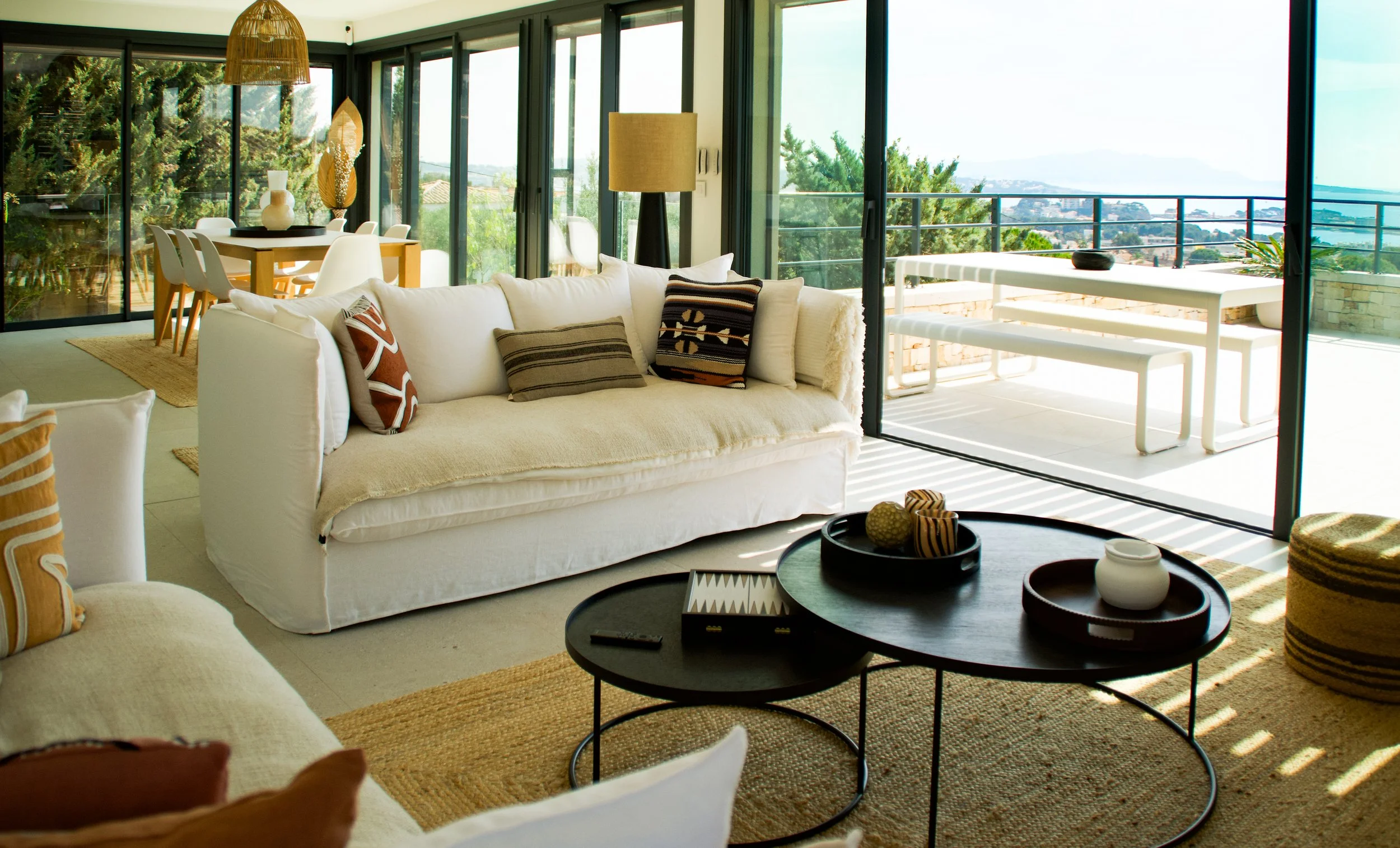 Photographie AirBnB Immobilier. Salon avec canapé blanc, tables basses rondes en bois, et grande baie vitrée ouverte sur un balcon avec vue sur la mer et la ville.