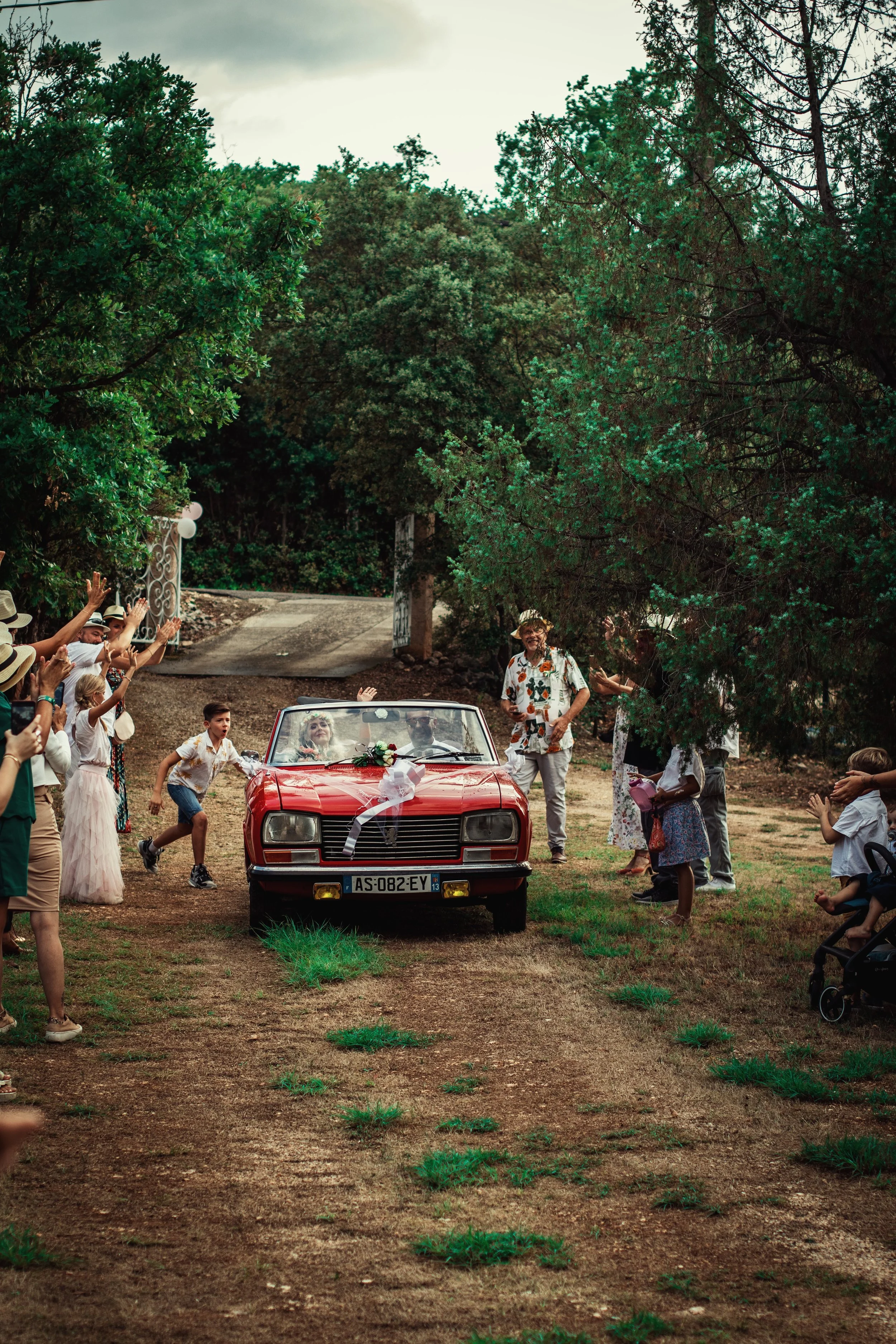 Photographe mariage - Une voiture de couleur rouge décorée pour un mariage, entourée d'invités applaudissant et célébrant dans un environnement naturel avec des arbres.