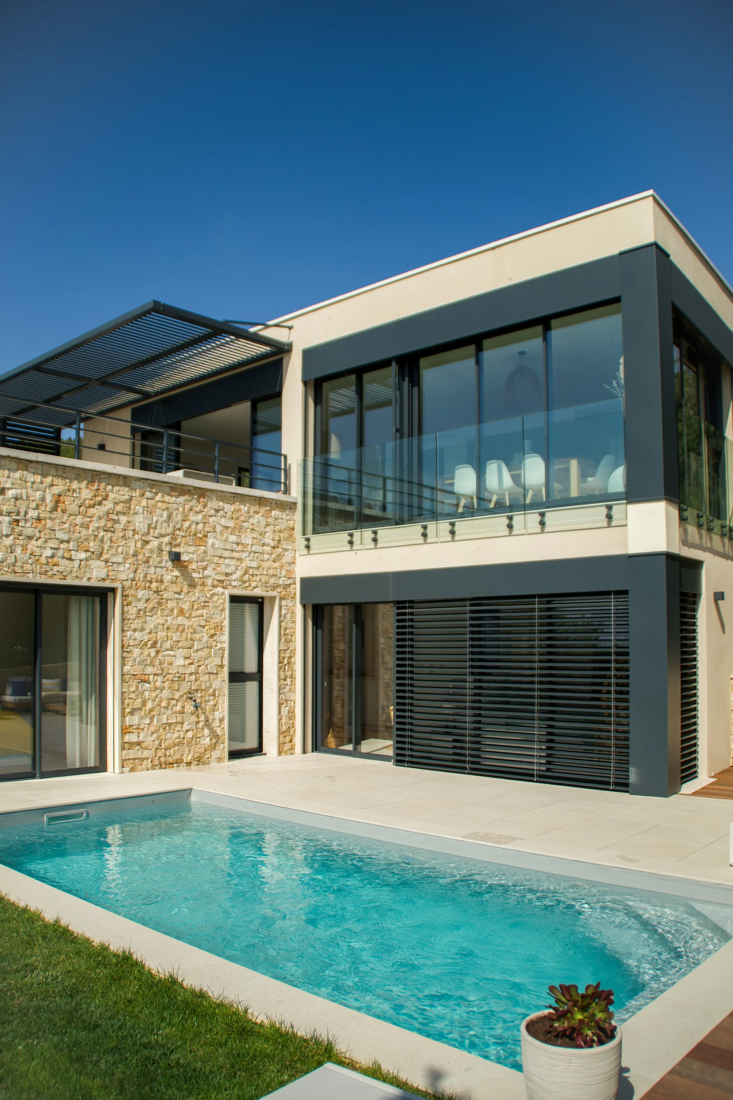 Photographie AirBnB Immobilier. Maison moderne avec piscine extérieure, façade en pierre et balcon en verre, sous un ciel bleu.