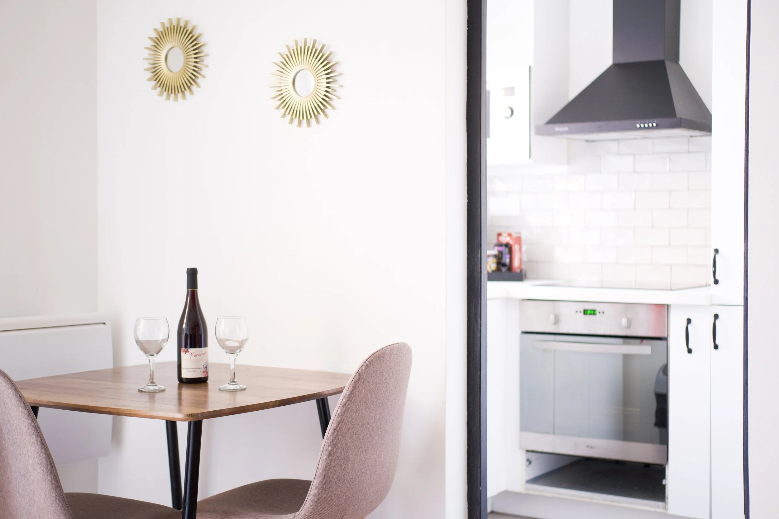 Photographie AirBnB Immobilier. Salle à manger avec une bouteille de vin et deux verres, cuisine en arrière-plan avec four et hotte aspirante, décoration murale en forme de soleil