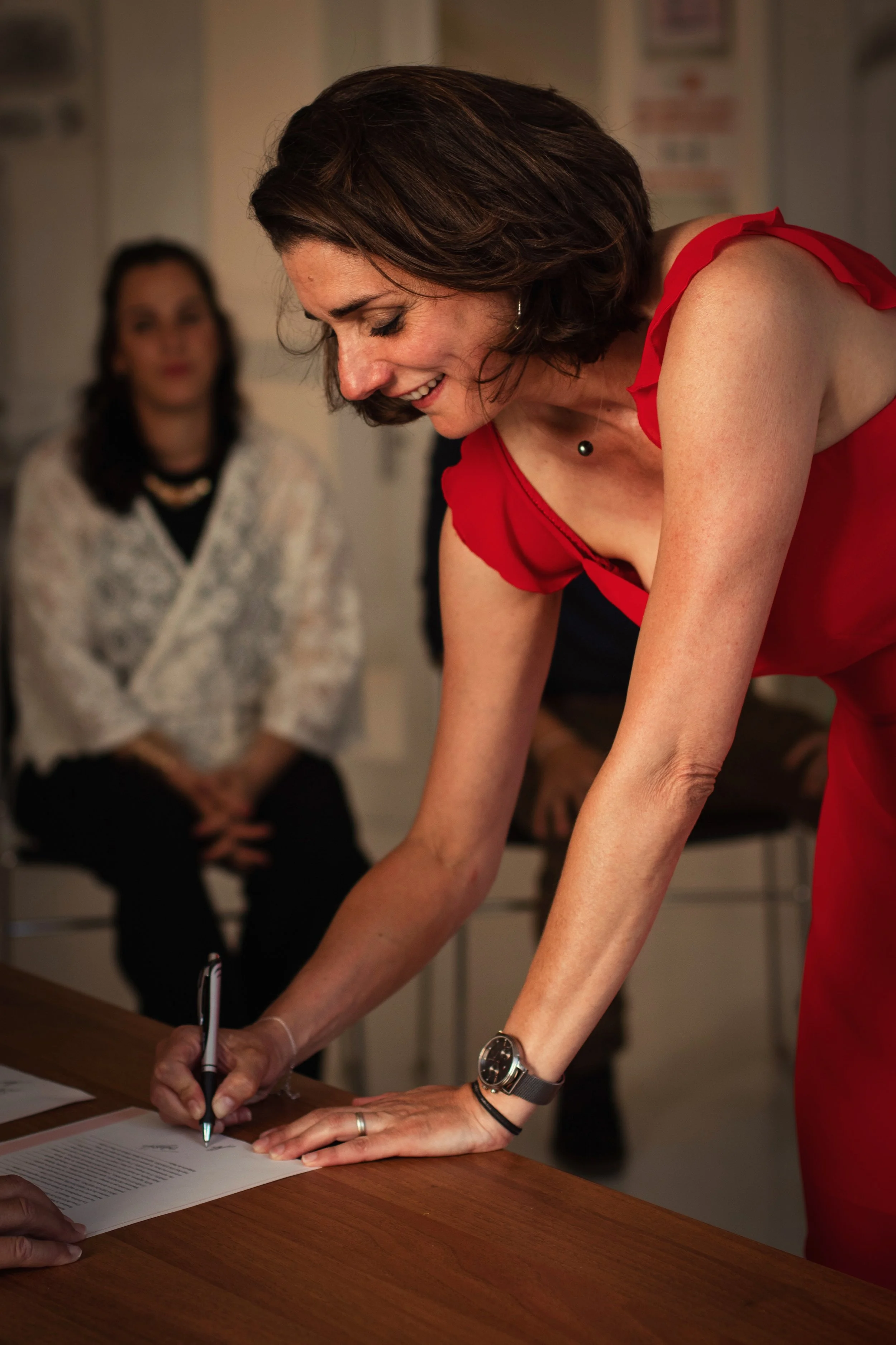 Photographe mariage - Une femme en robe rouge signant un document sur une table, avec deux femmes en arrière-plan assises, dans un cadre intérieur.