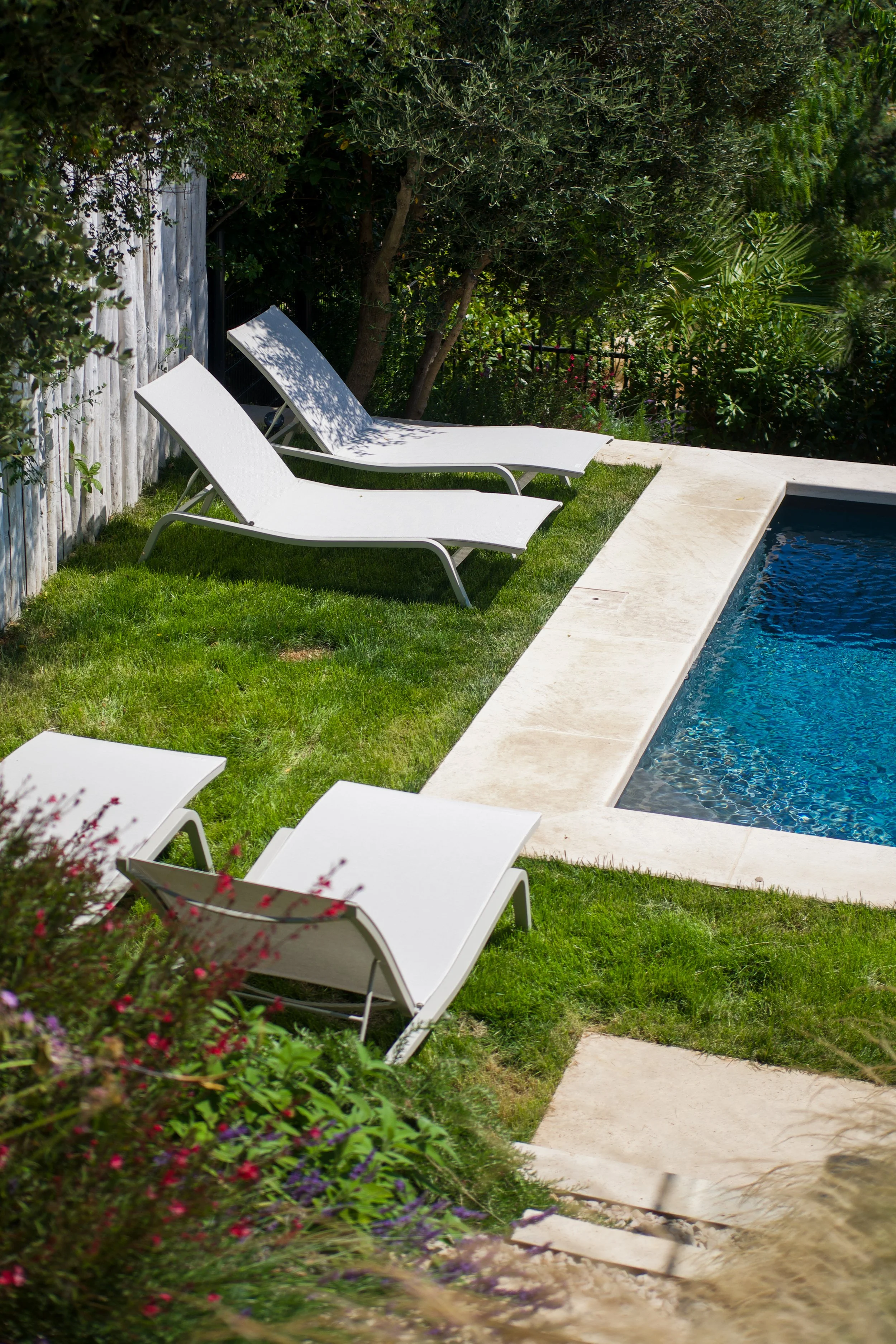 Photographie AirBnB Immobilier. Deux chaises longues blanches au bord d'une piscine, entourées de verdure et de fleurs. 