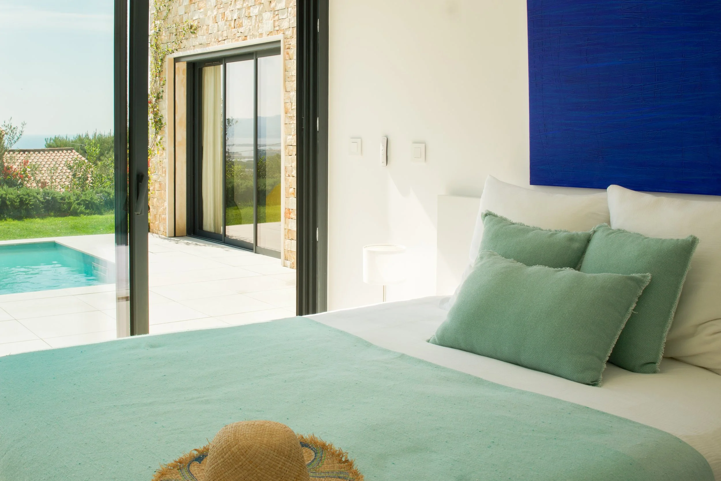 Photographie AirBnB Immobilier. Chambre avec lit blanc et coussins verts, vue sur une terrasse extérieure avec piscine, au-delà de jardins verdoyants et maison avec toiture en tuiles.