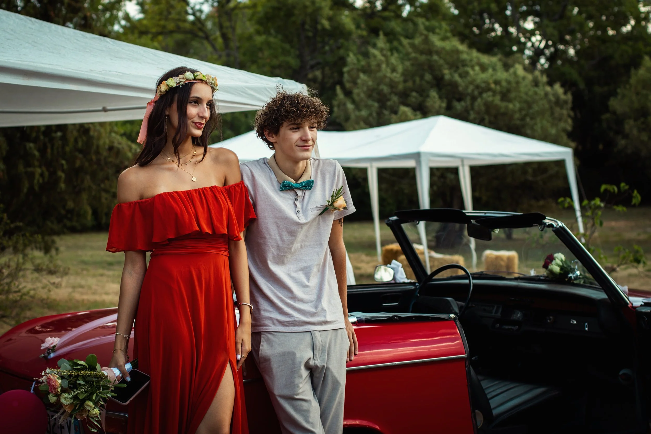 Photographe mariage - Deux jeun assis sur une voiture rouge lors d'une fête en plein air. La femme porte une robe rouge et une couronne de fleurs. L'homme porte une chemise claire avec un nœud papillon vert. Deux tentes blanches et une forêt en fond.