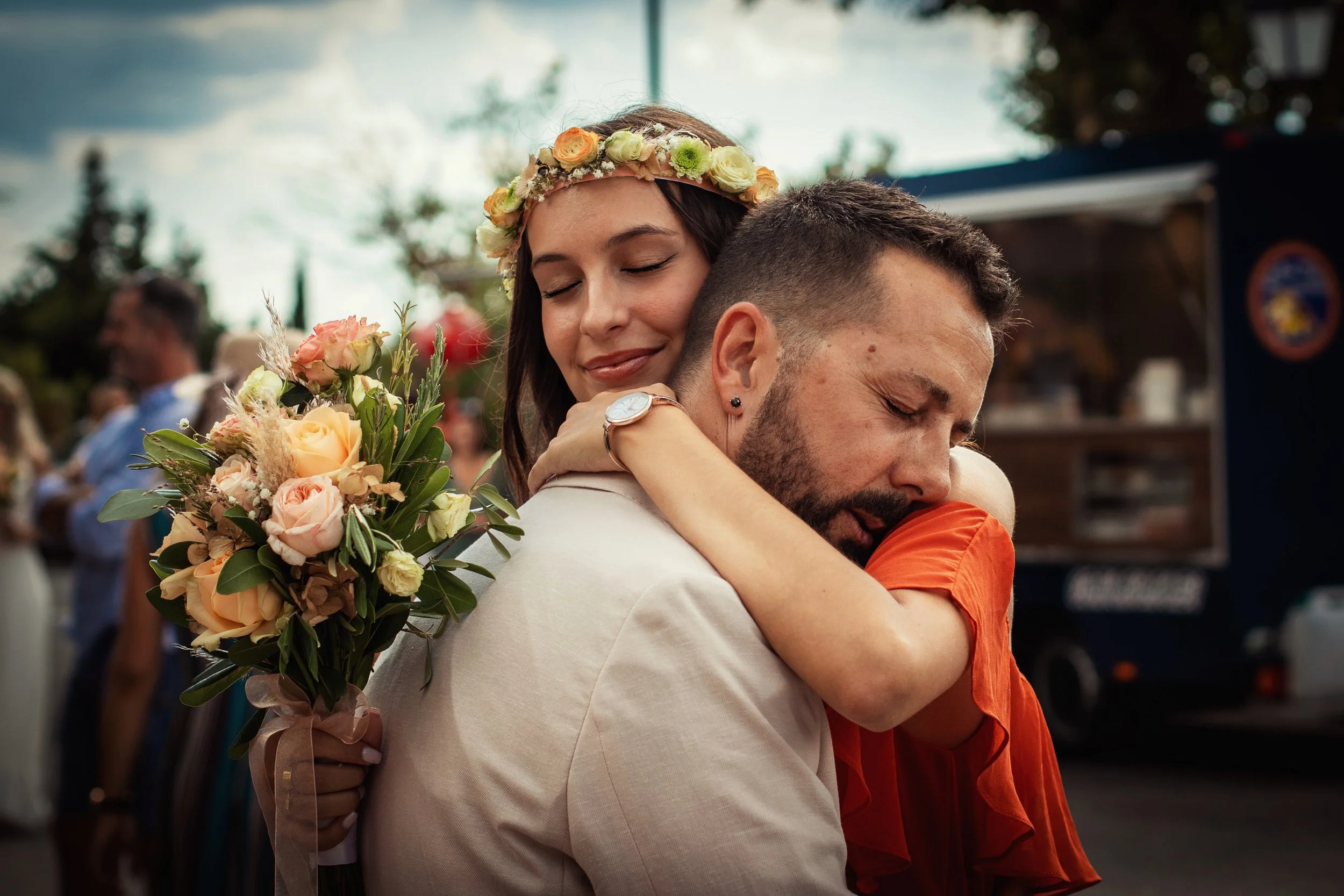 Photographe mariage - Deux personnes s'étreignant chaleureusement à une occasion spéciale, une femme porte une couronne de fleurs et tient un bouquet de fleurs, dans un environnement extérieur avec d'autres personnes en arrière-plan.