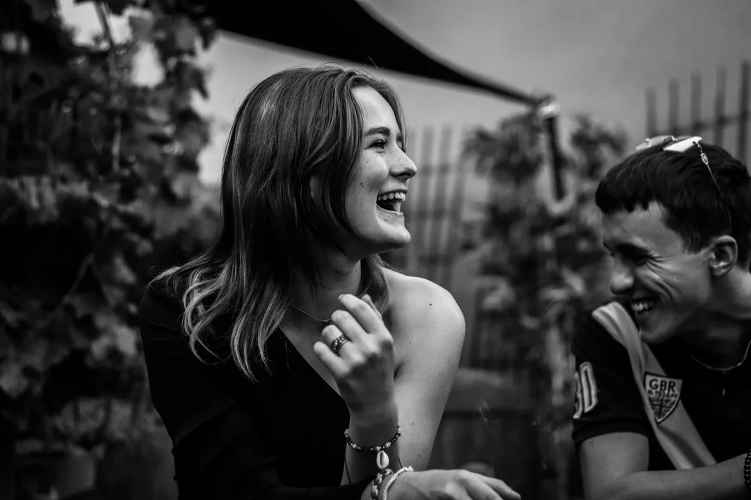 Photographe événement - Deux jeunes personnes souriantes qui rient, une femme avec des cheveux mi-longs bruns et un homme portant des lunettes de soleil, dans un environnement extérieur.