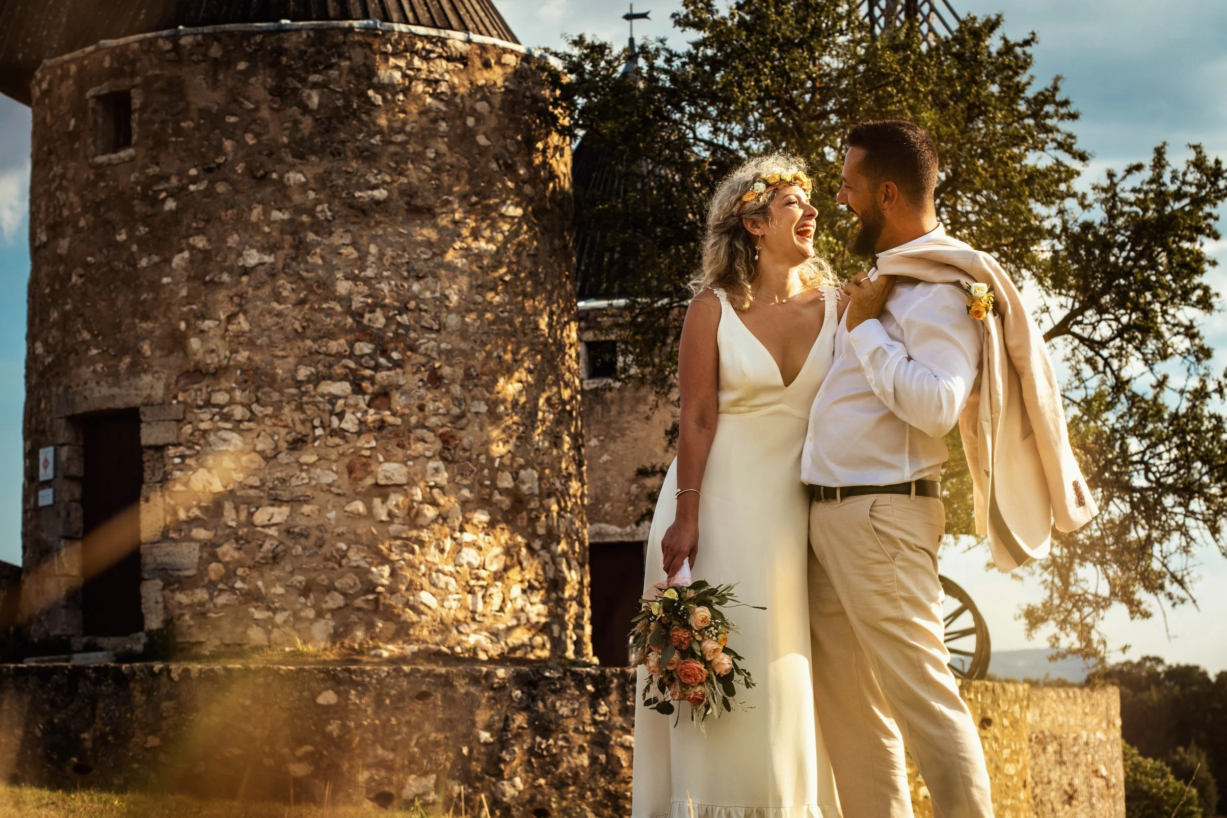 Photographe mariage - Un couple de mariage heureux souriant à l'extérieur, avec un vieux moulin en pierre en arrière-plan, entouré d'arbres, lors d'une journée ensoleillée.