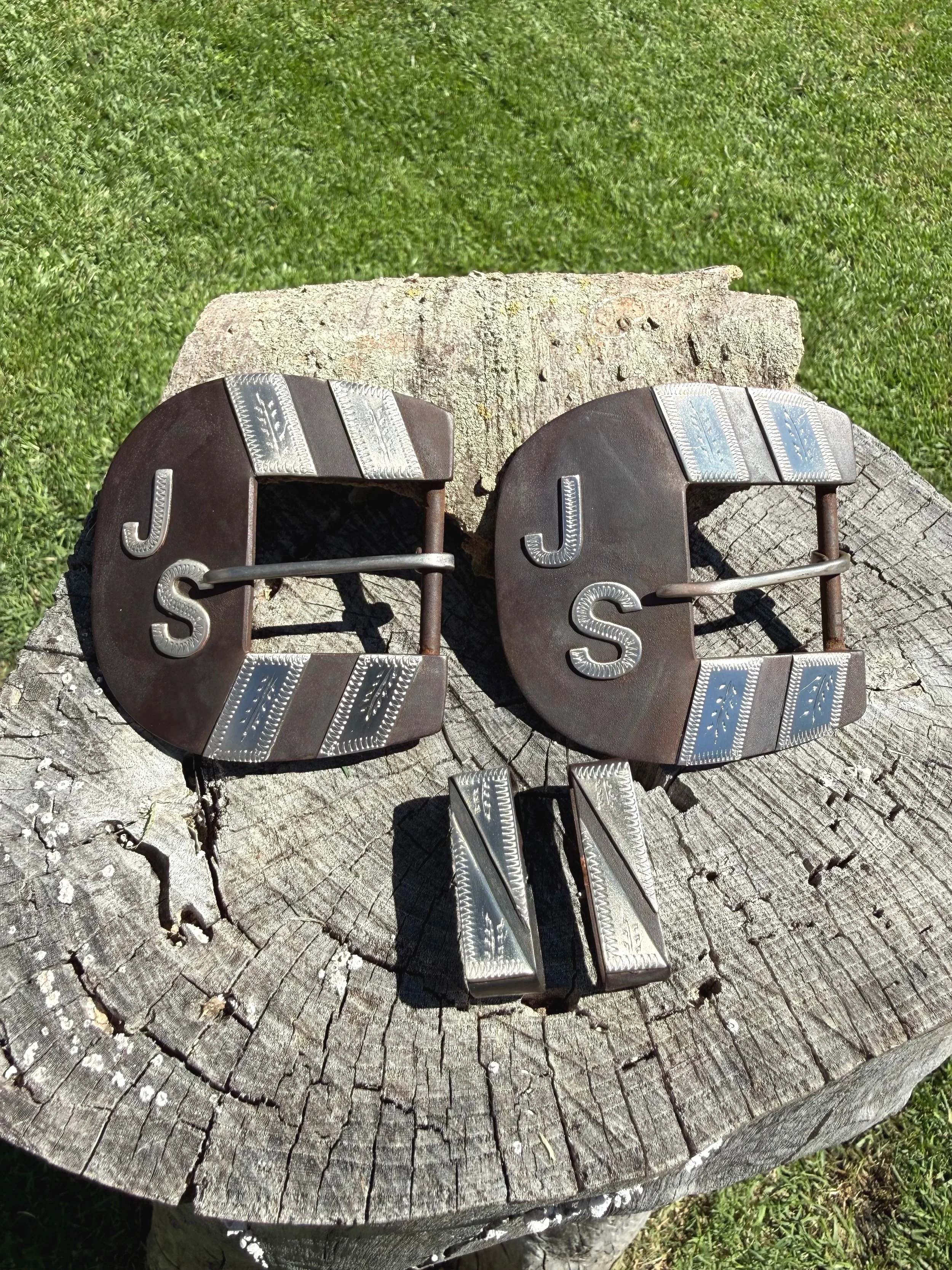 A pair of Western-style belt buckles with silver embellishments, placed on a weathered tree stump outdoors.