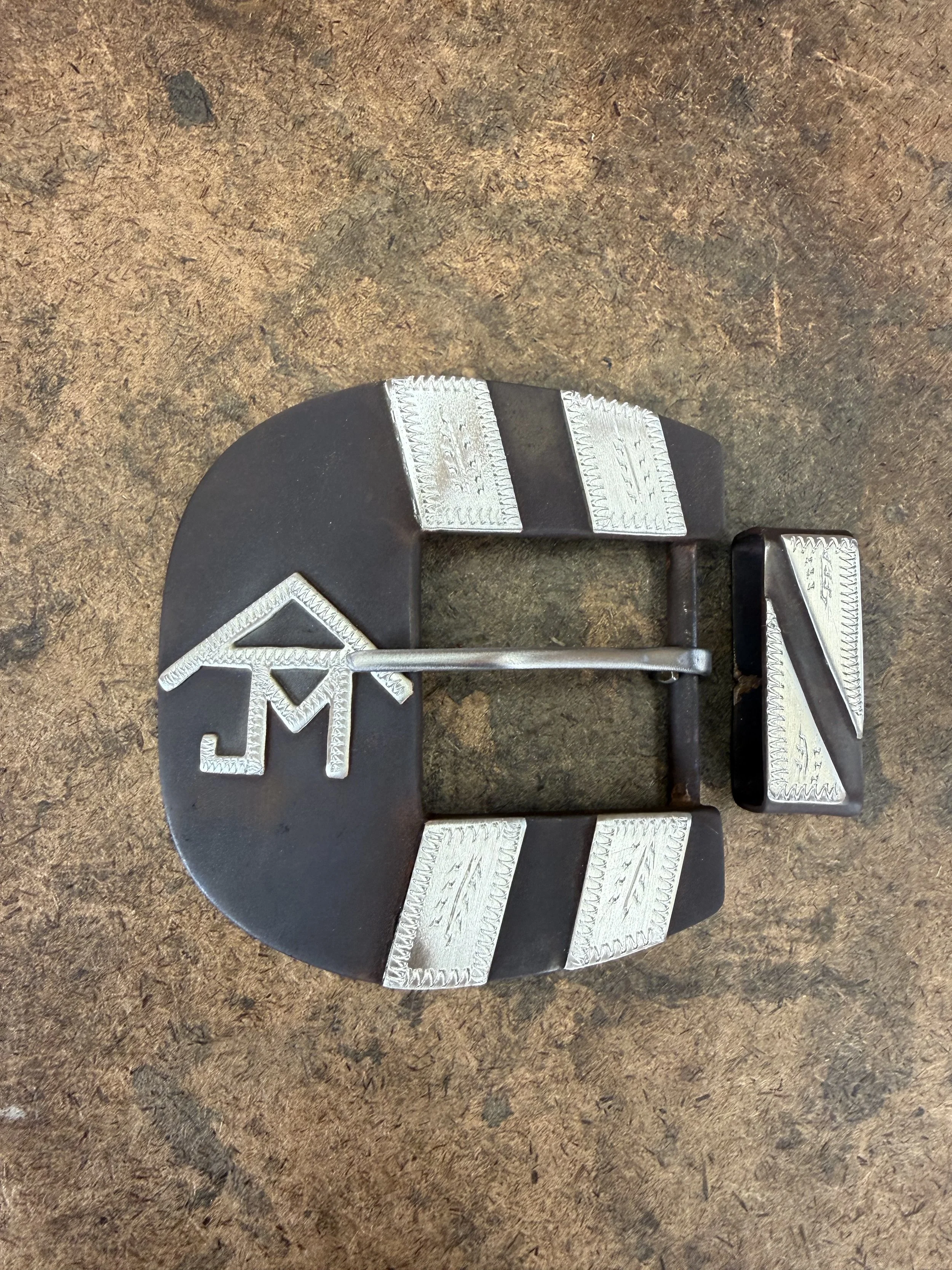 A circular black exercise weight plate with silver metal strips and attached weightlifting hooks, placed on a brown textured surface.