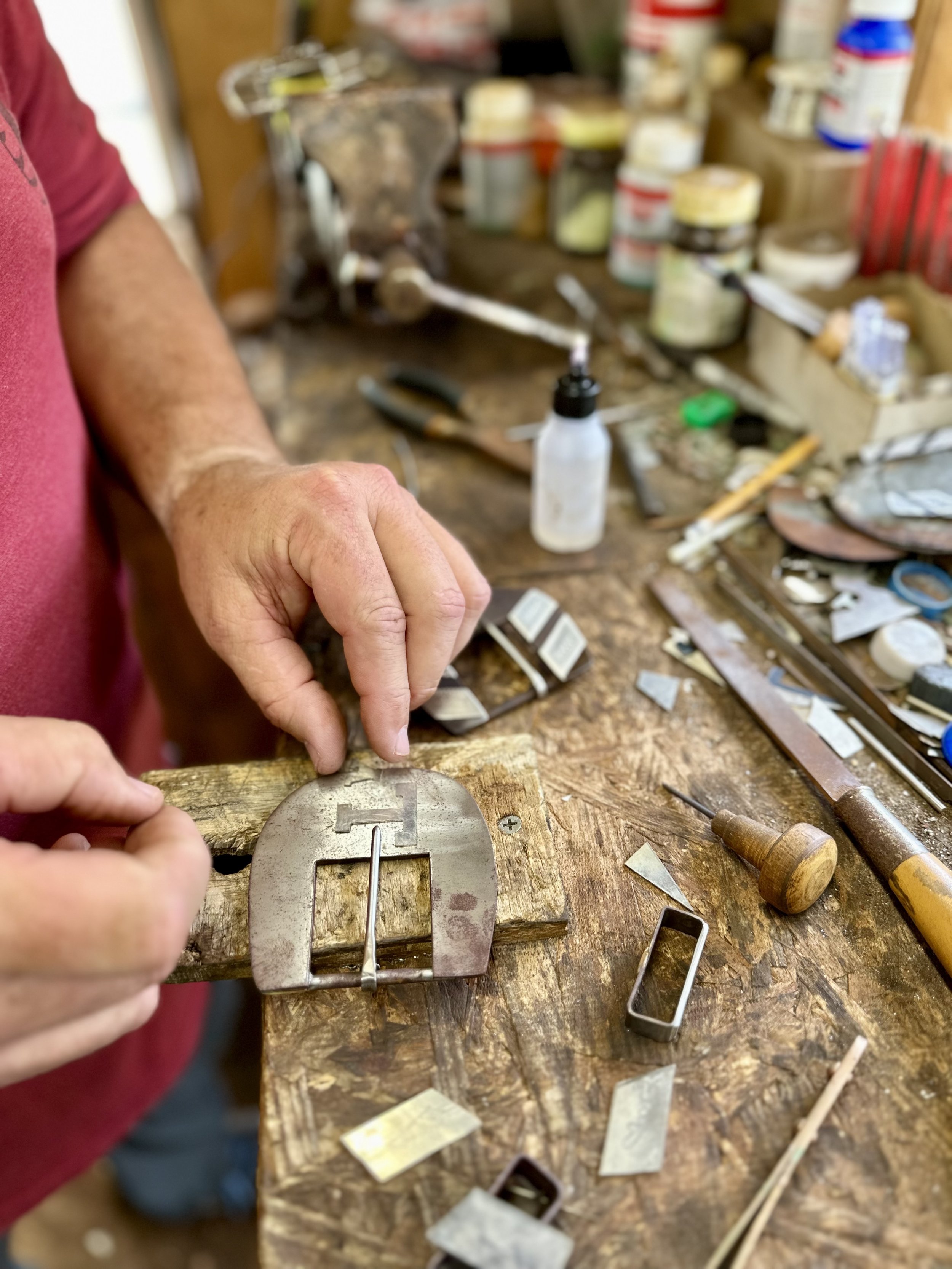 Person working on a craft or jewelry making project at a cluttered workbench with various tools and materials.