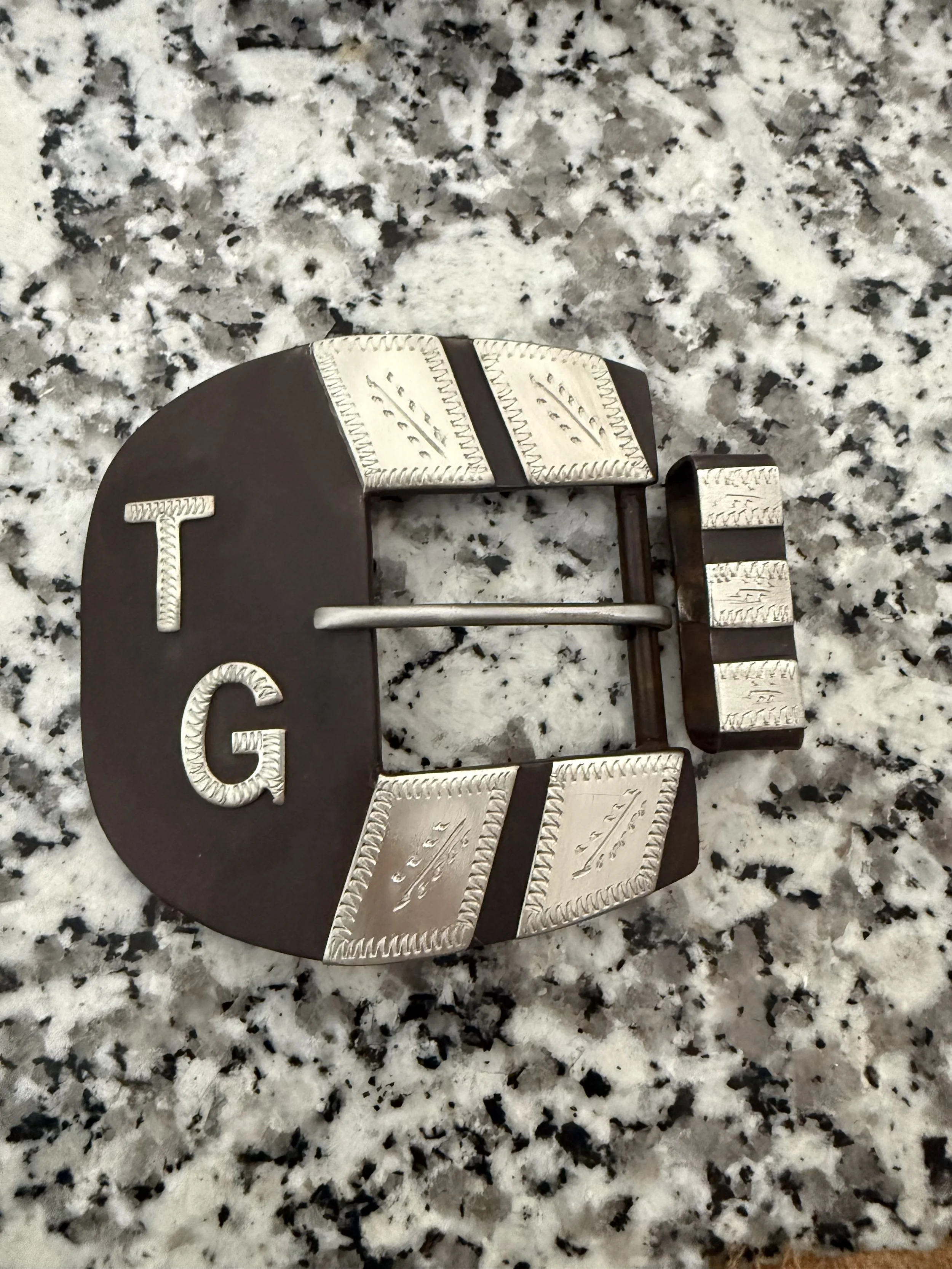 Close-up of a decorative metal sign on a granite countertop. The sign has the word 'WELCOME' spelled out with individual letters, and the background surface is speckled with black, white, and gray colors.