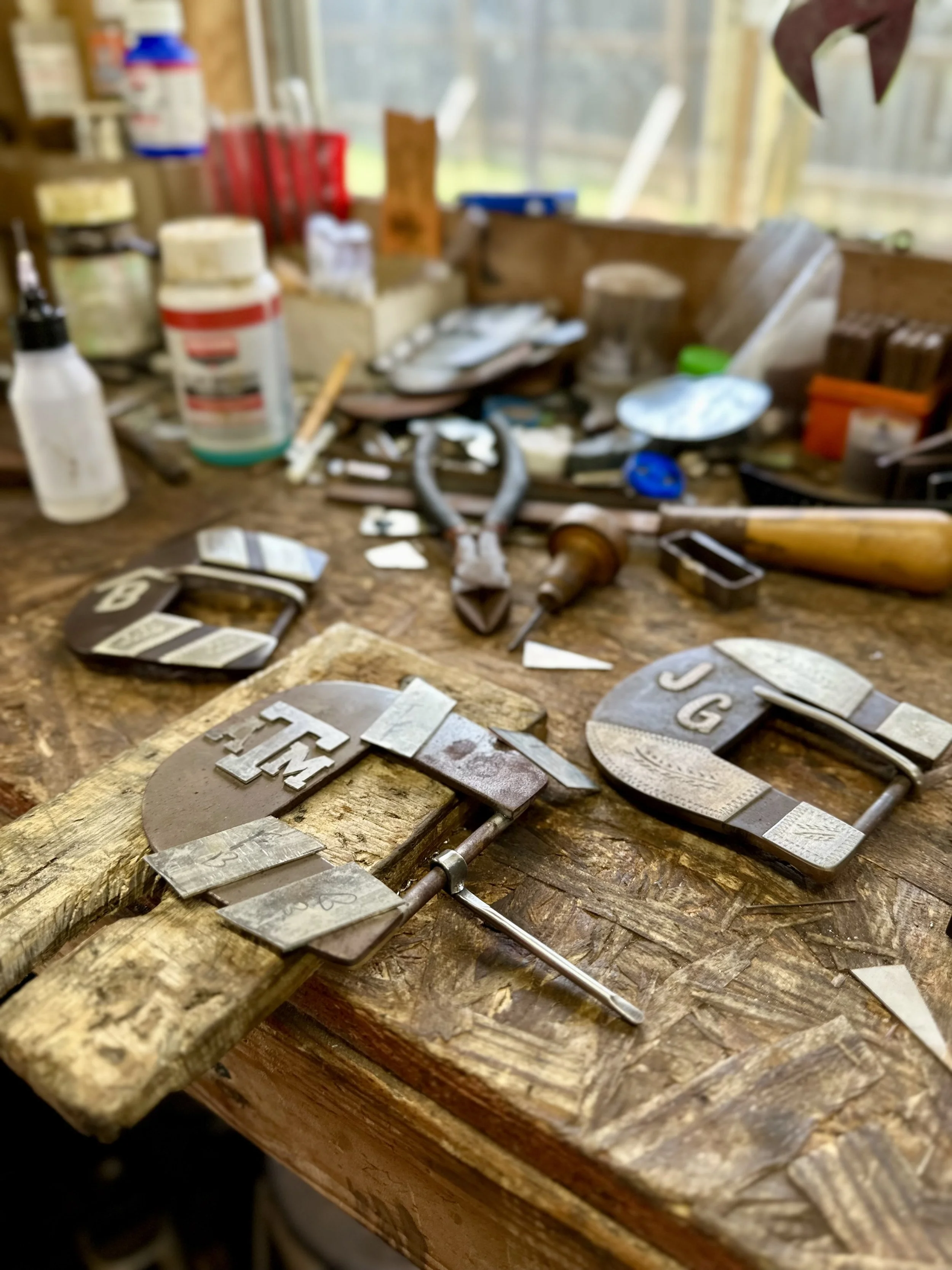 Close-up view of a cluttered workshop table with metal jewelry letters and tools, with a blurry background of workshop supplies.