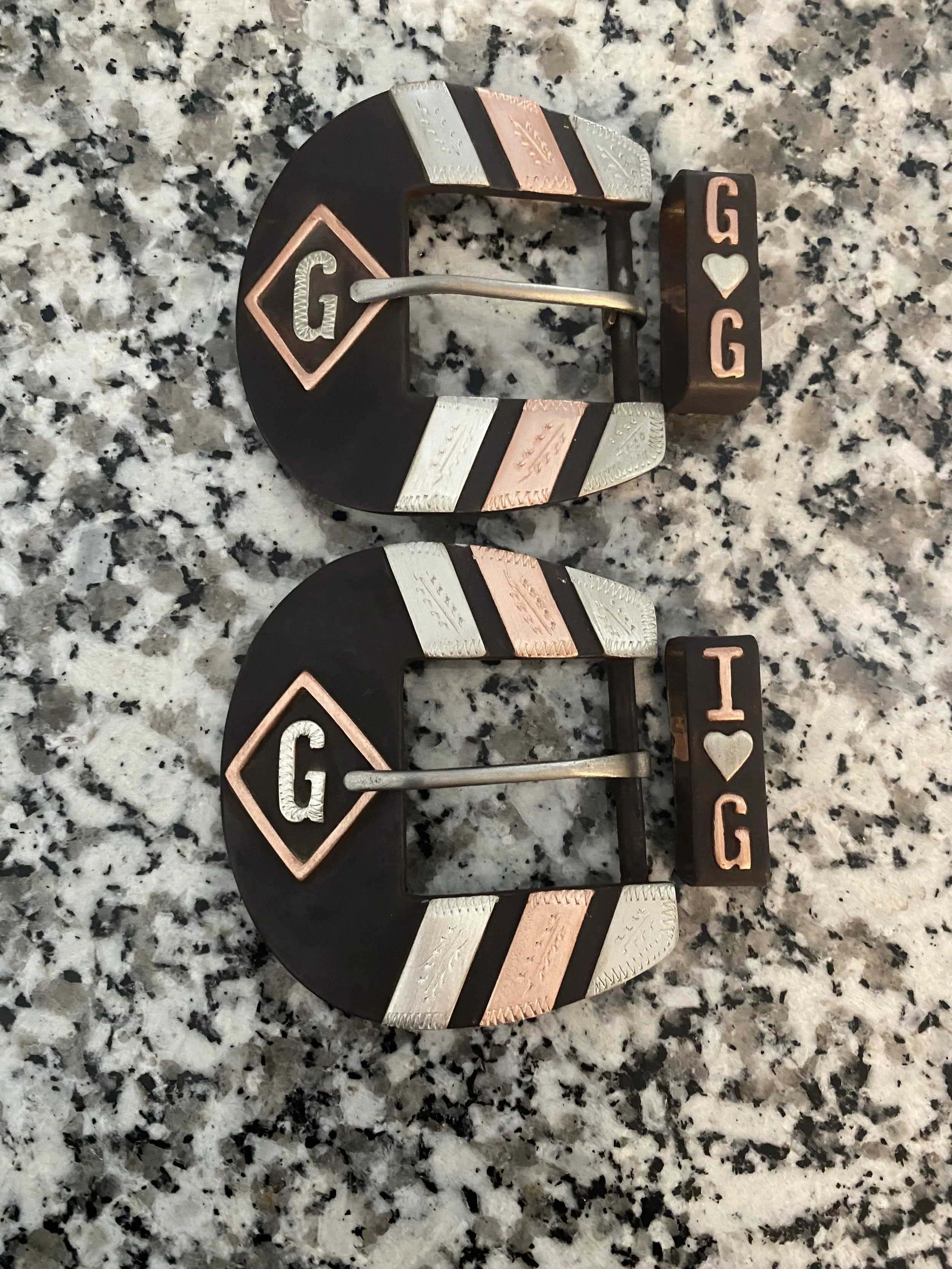Two belt buckles with the phrase "G ♥ G" on a speckled granite countertop.