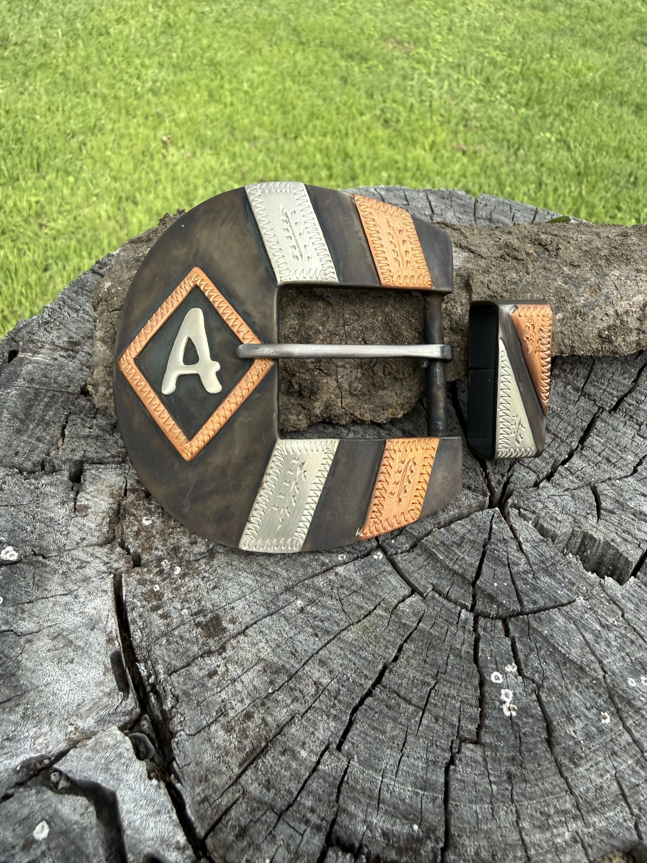 A decorative belt buckle with an orange, silver, and black design, with the letter 'A' in the center, resting on a weathered, cracked piece of wood with green grass in the background.