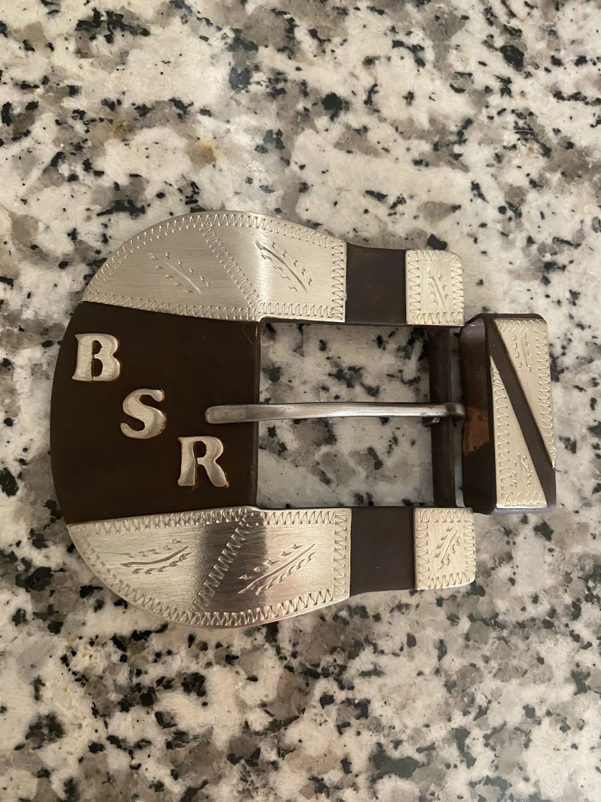 Close-up of a metallic belt buckle with engraved decorative details and the letters 'BS R' on a speckled granite countertop.