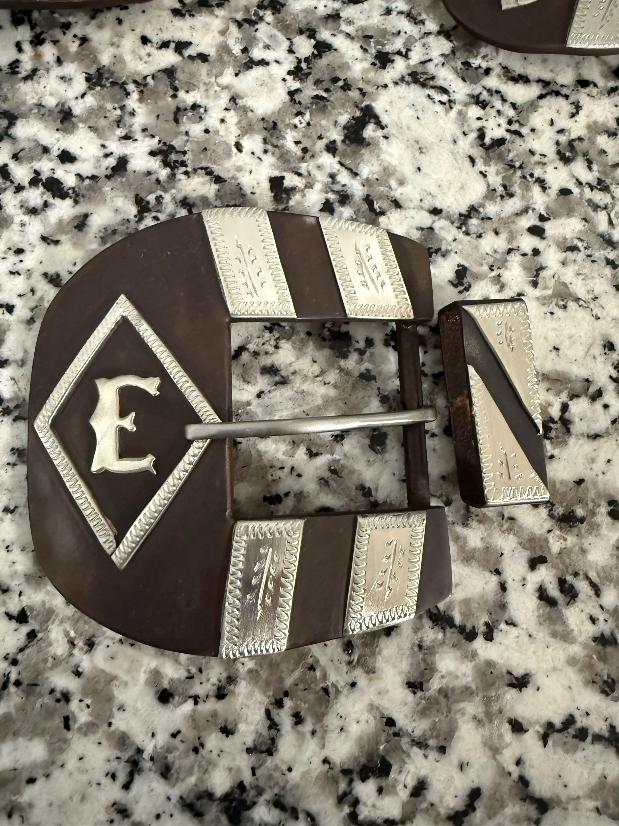 Close-up of a chocolate cake decorated with silver edible accents and a silver letter 'E' on a silver square, placed on a speckled granite surface.