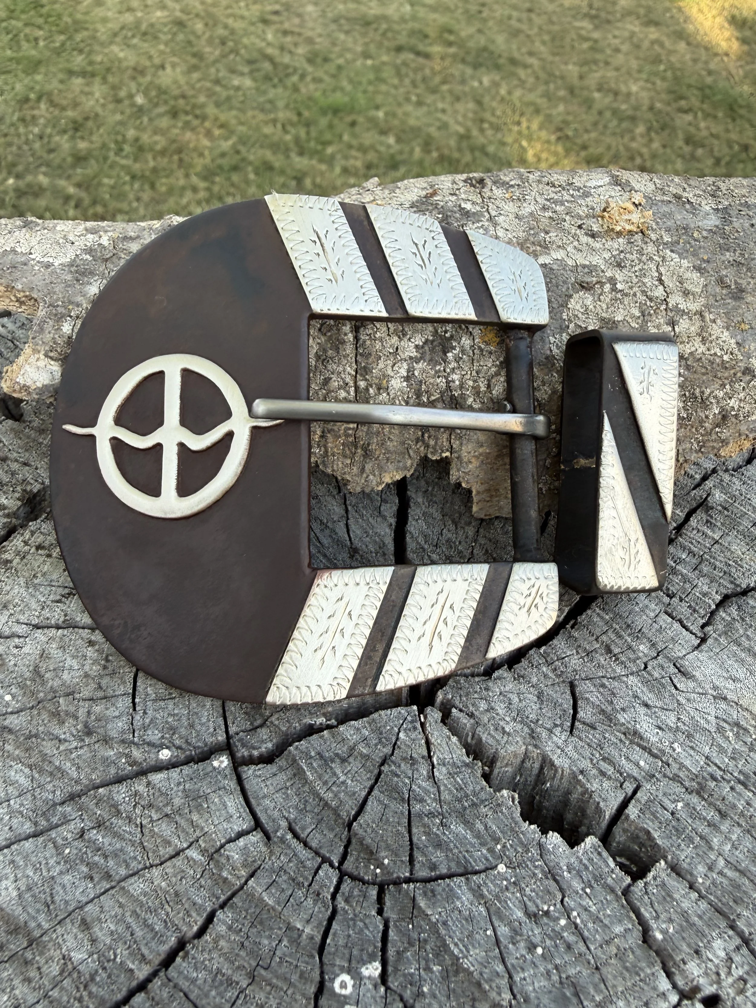 A vintage black and silver belt buckle with a peace sign symbol, placed on a tree stump with cracks and rings.