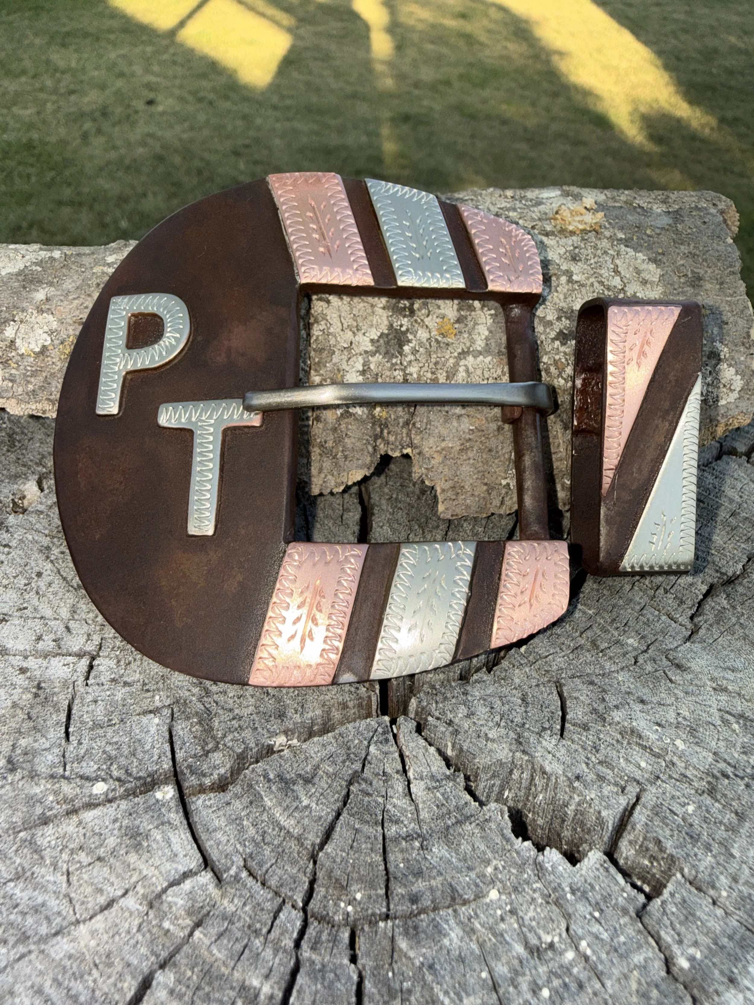 A decorative belt with the letters P, F, and V, placed on a cracked wooden surface outdoors with grass and shadows in the background.