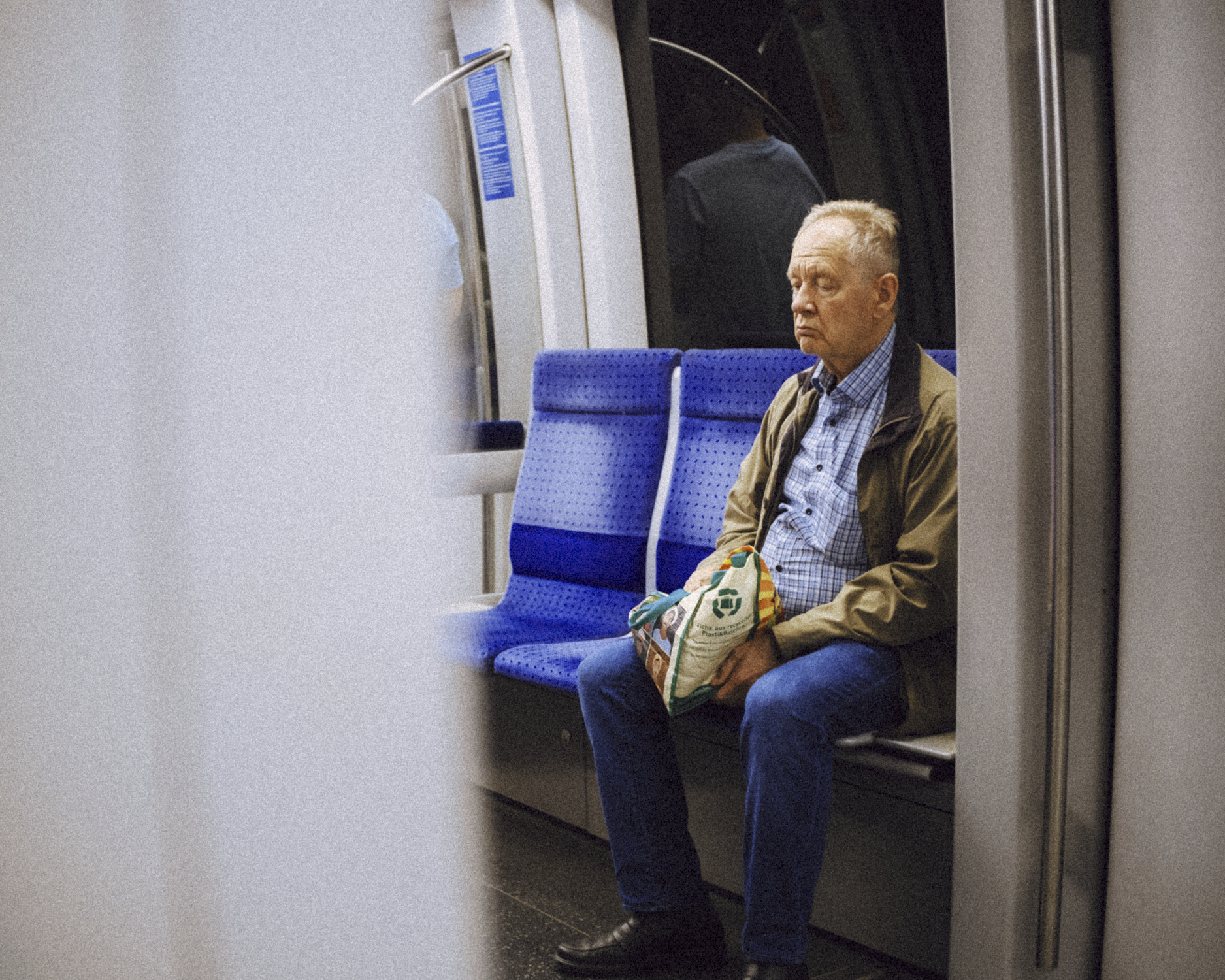 An older man sitting alone on a blue seat on a subway train, holding a grocery bag, with a reflective window behind him and a partial view of another passenger in the background.