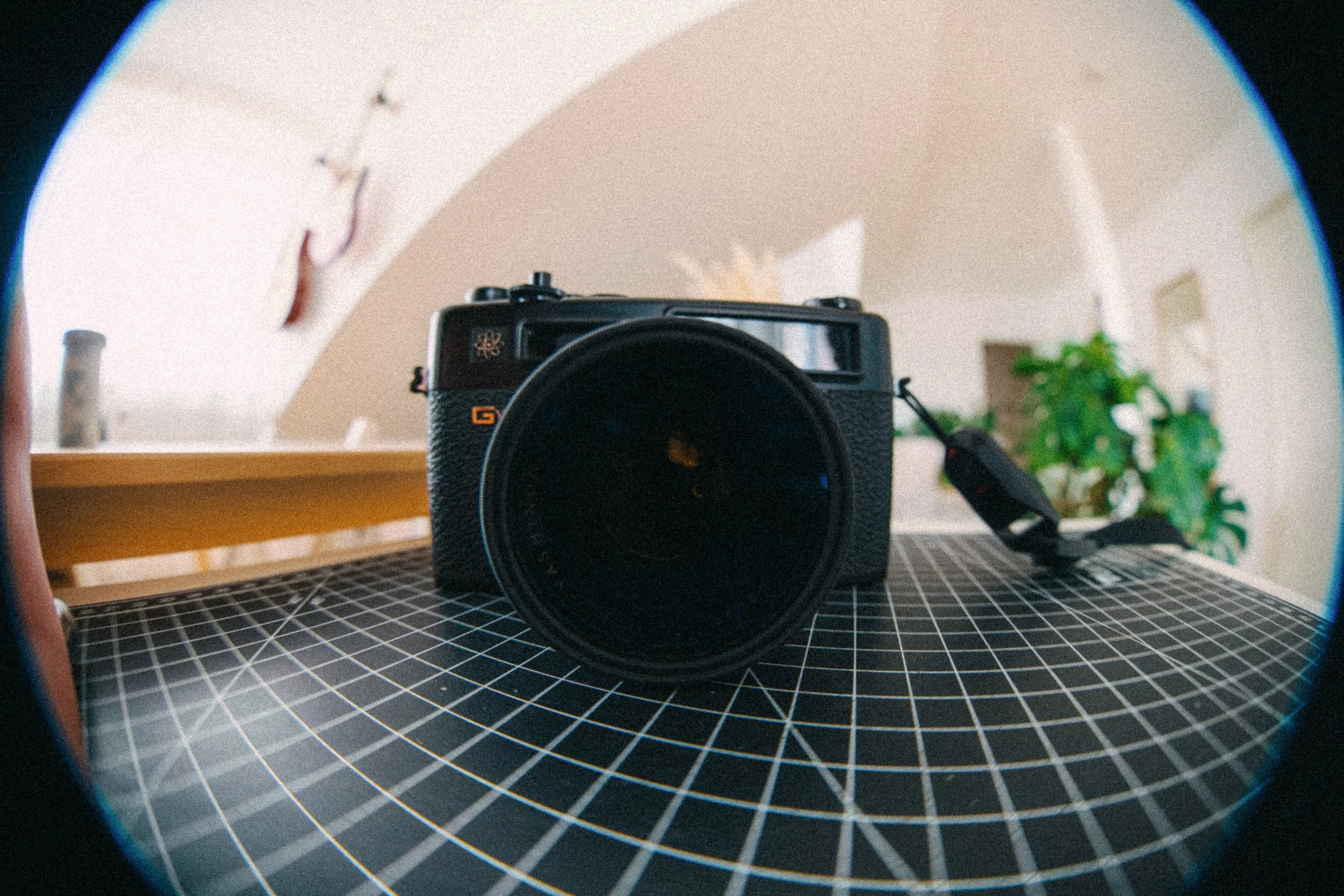 A camera with a large lens, placed on a black grid-patterned surface, taken with a fisheye lens, showing a room with a wooden table and a green plant in the background.