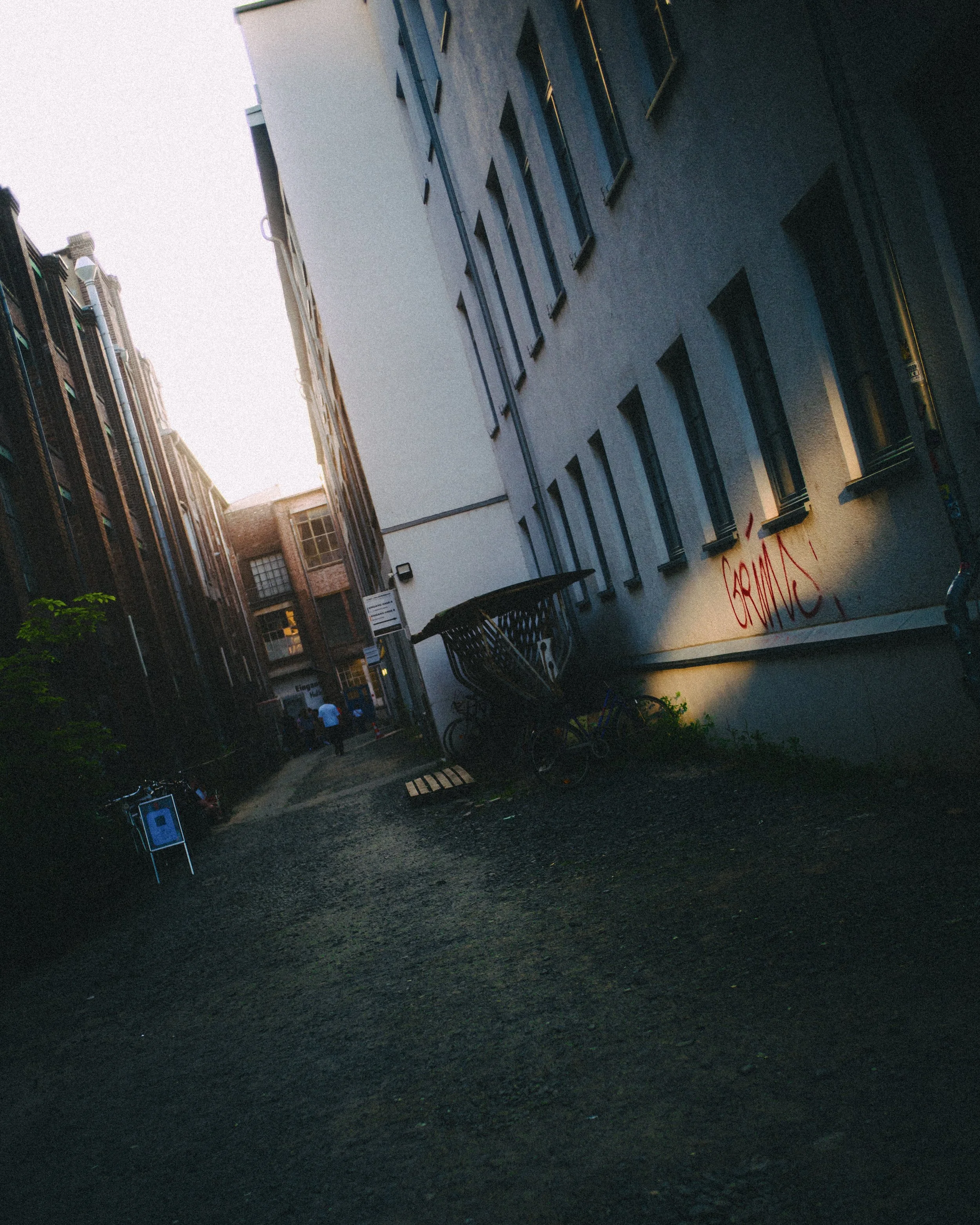 An alleyway between tall buildings with a cart, graffiti on the wall, and a few people in the distance at dusk.