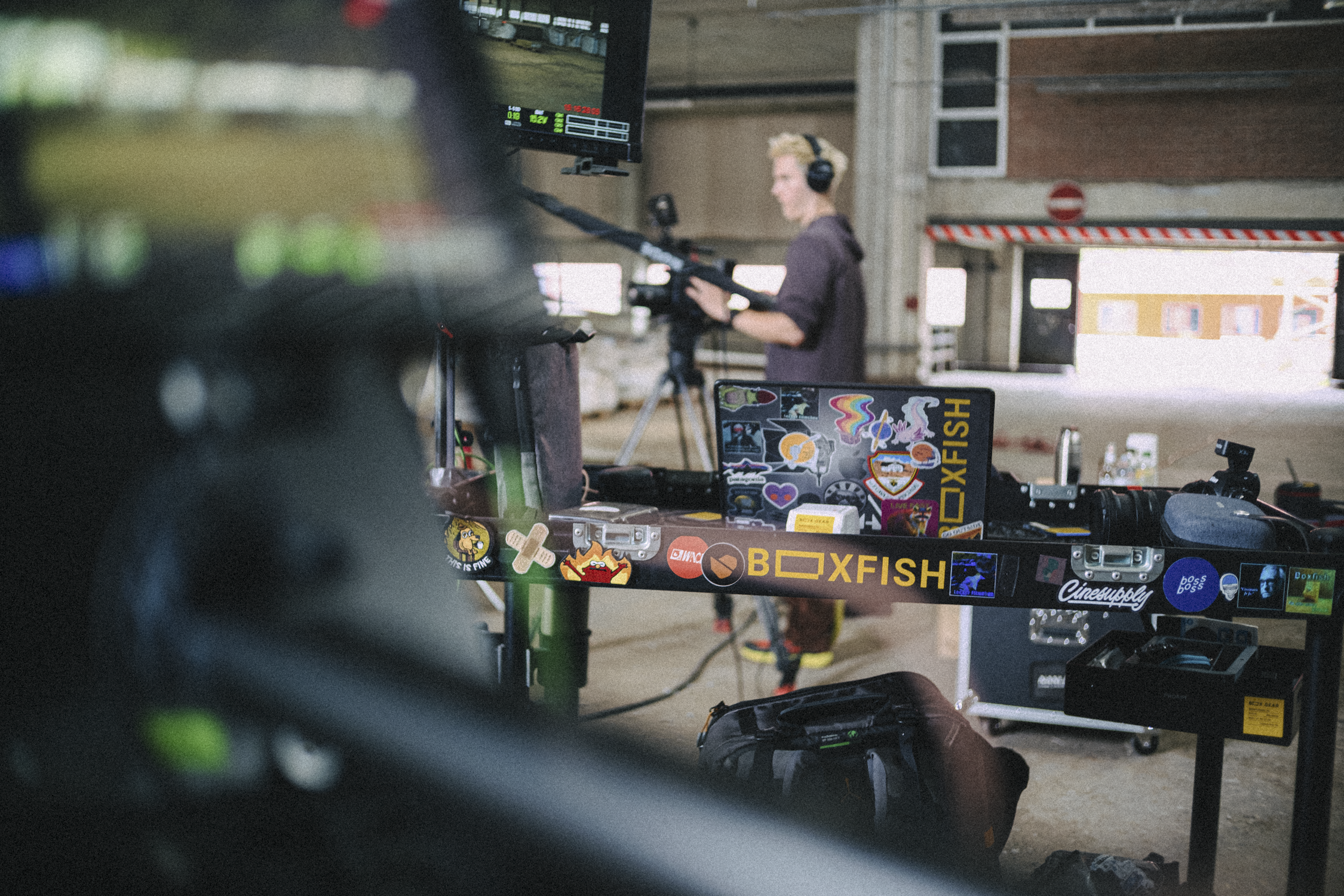 A person in a dark shirt and headphones operates a camera on a tripod inside an industrial or warehouse-like space, with various equipment and a monitor visible.