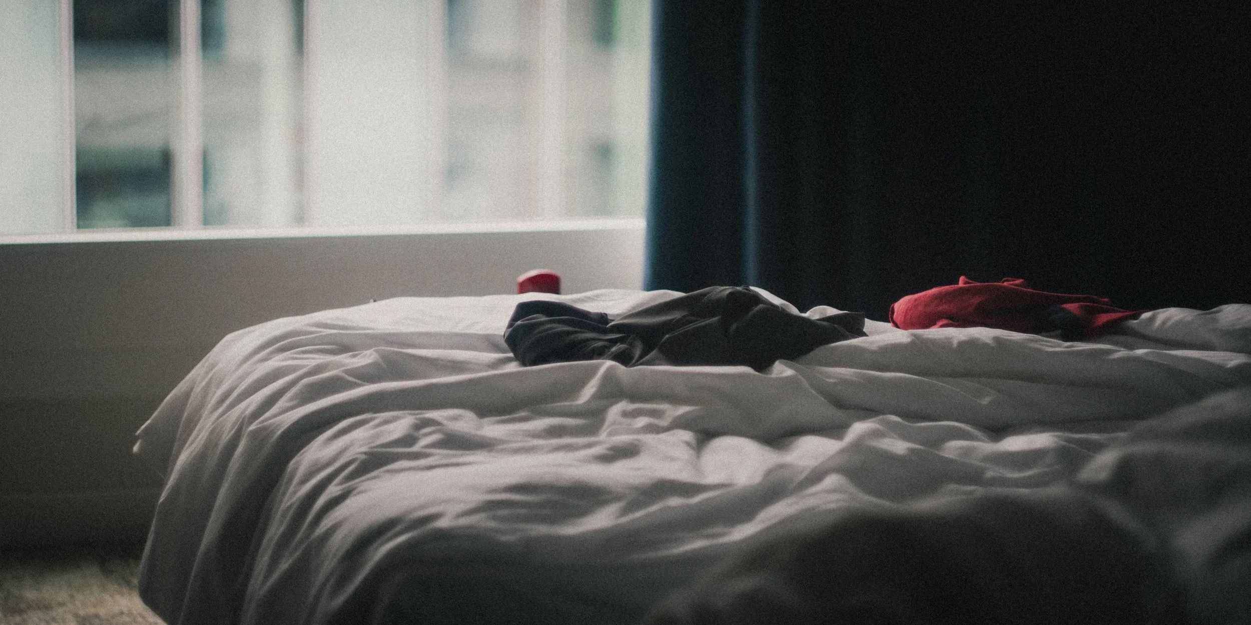 A messy bed with crumpled white sheets, a navy sock, and a red cloth near a large window with a sheer curtain, in a dimly lit room.
