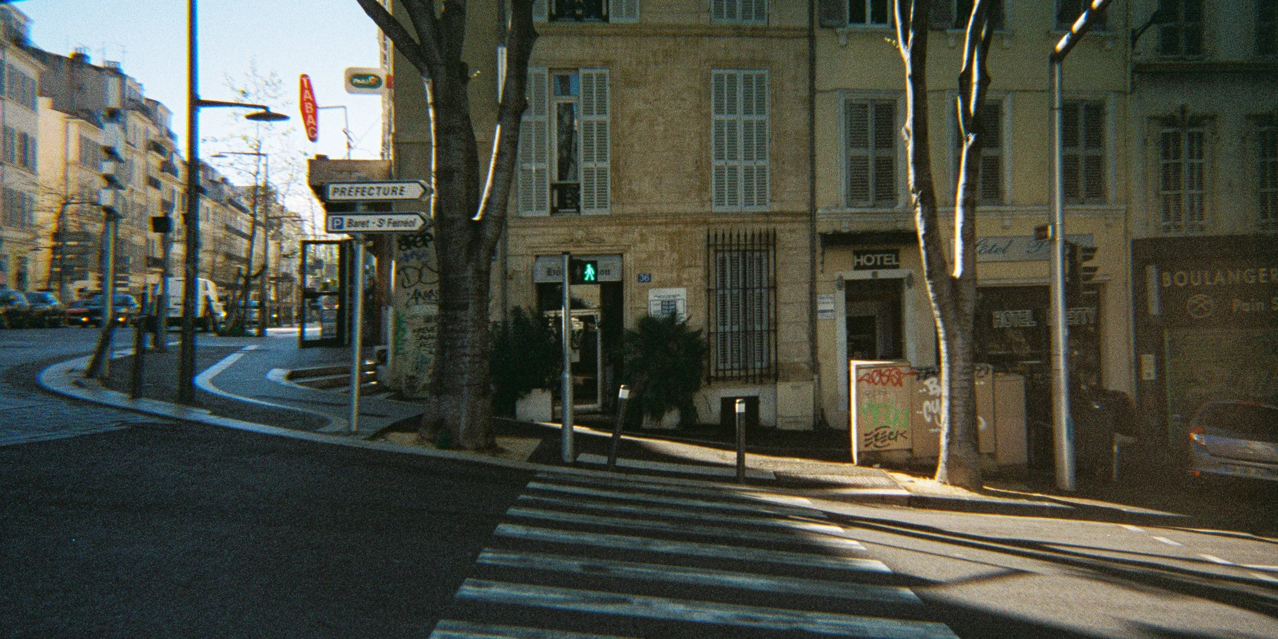 City street scene with a pedestrian traffic light, buildings, trees, a hotel, graffiti on a sign, parked cars, and a crosswalk during daytime with sunlight and shadows.