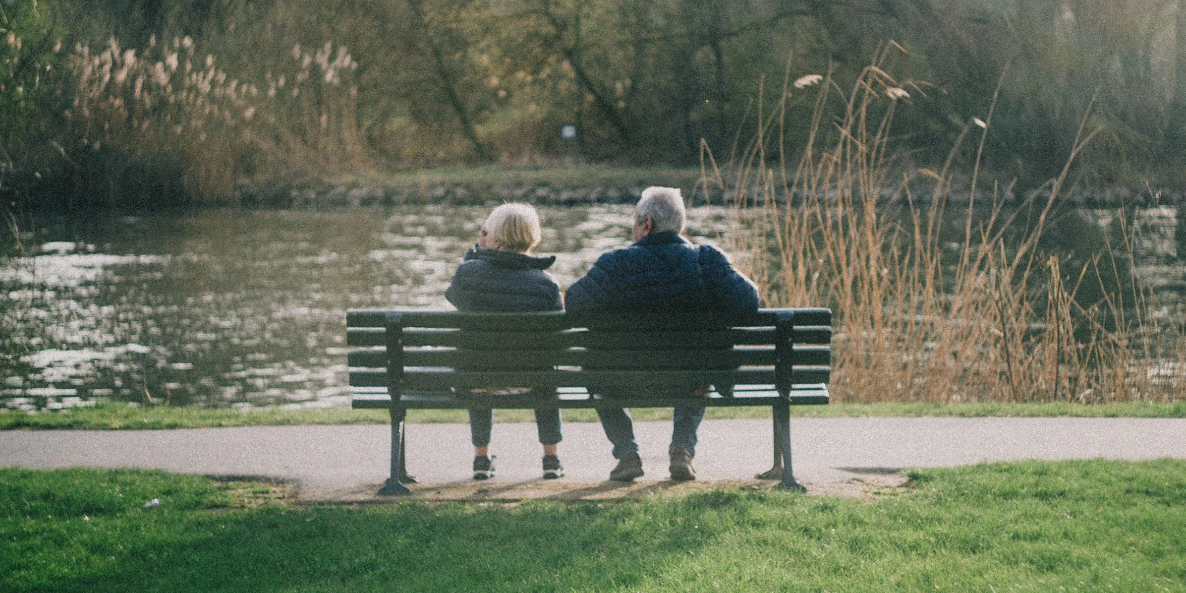 An elderly man and woman sitting close together on a park bench by a pond, engaging in conversation.