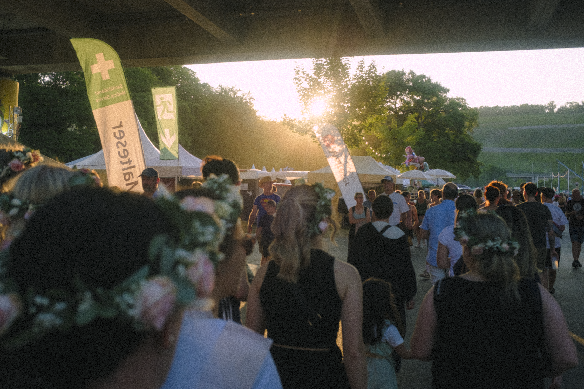 Crowd of people attending an outdoor event or festival during sunset, some wearing floral headbands, surrounded by tents and flags.