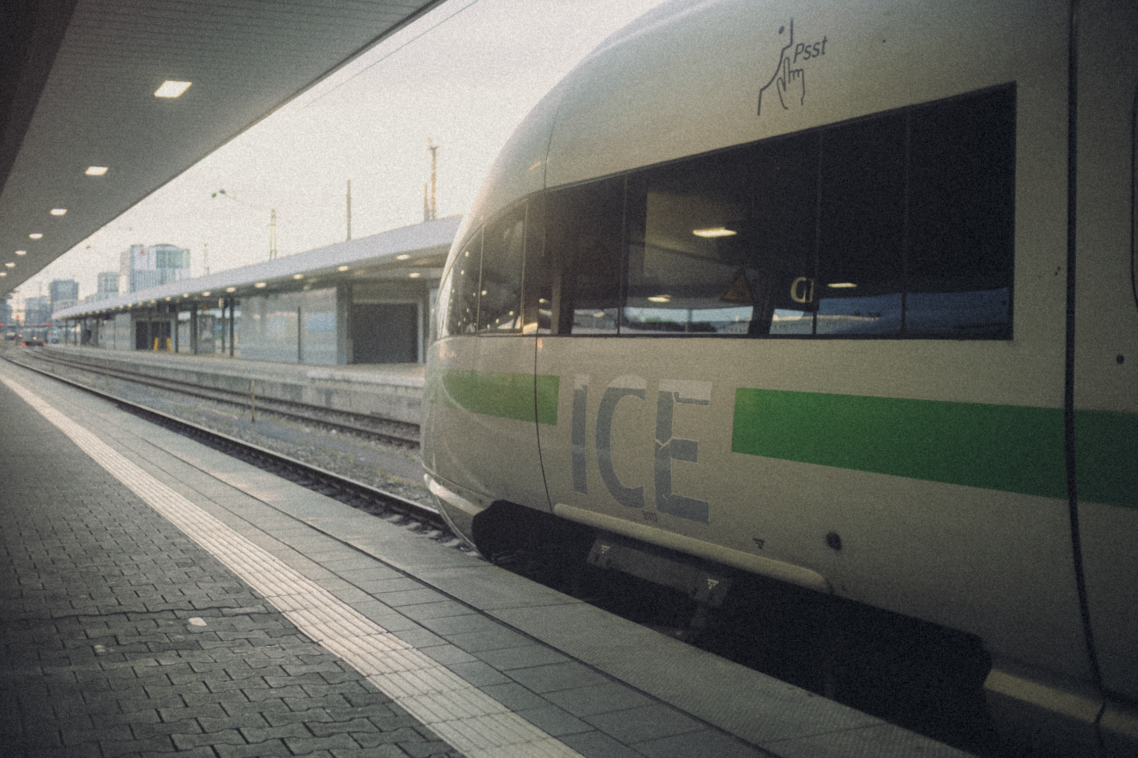 A modern train station platform with an ICE train on the right side. The train has large windows and the word 'ICE' visible on the side. The platform is empty, with a covered waiting area and buildings in the background, under a cloudy sky.