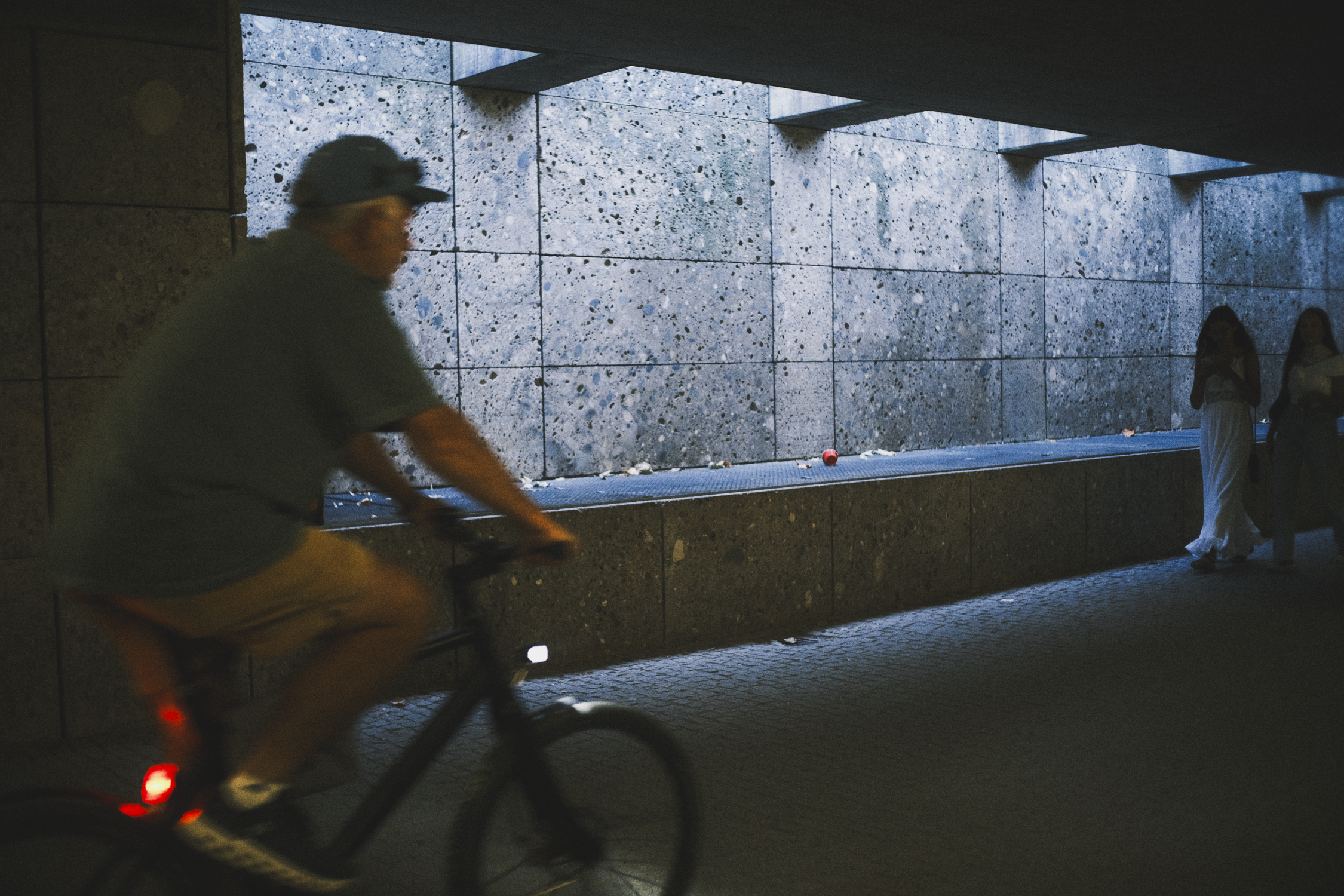 A person riding a bike past a wall with a ledge under a bridge, and two women standing nearby on the right.