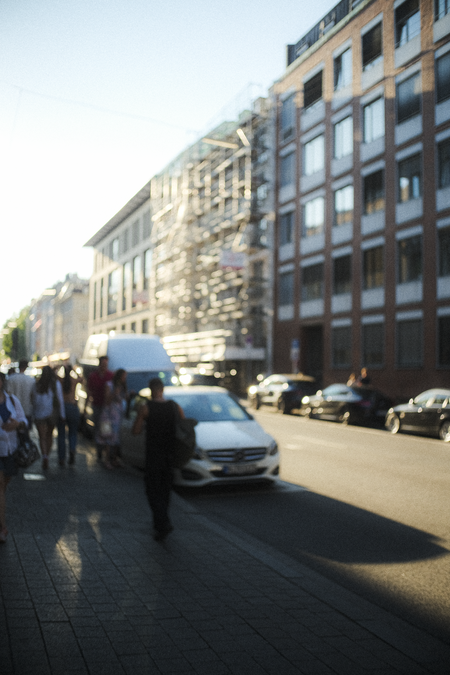 City street scene with pedestrians walking on the sidewalk, cars parked along the street, and multi-story buildings with modern architecture.