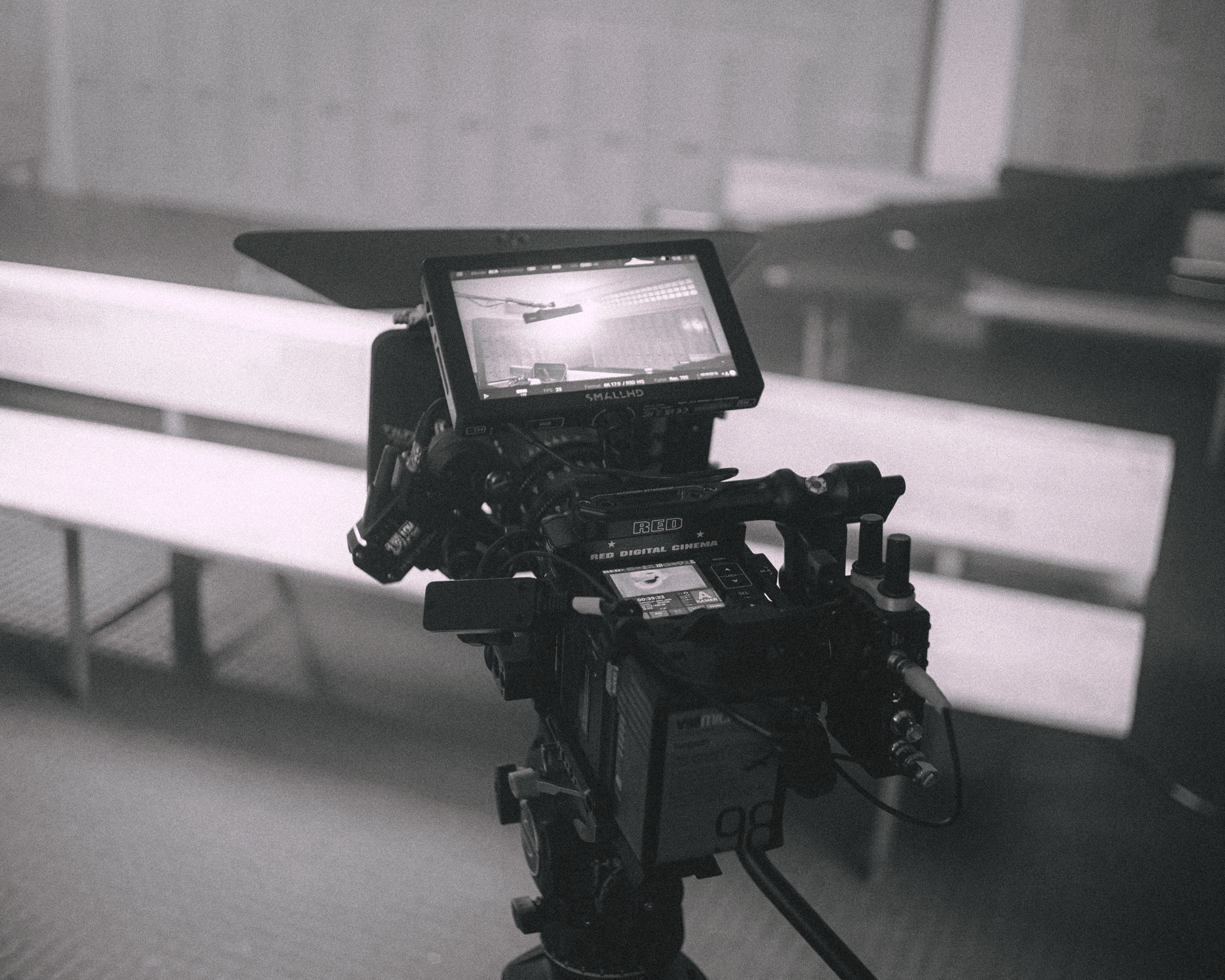 A professional video camera mounted on a tripod, with a monitor attached, filming a scene in an indoor setting with benches in the background.