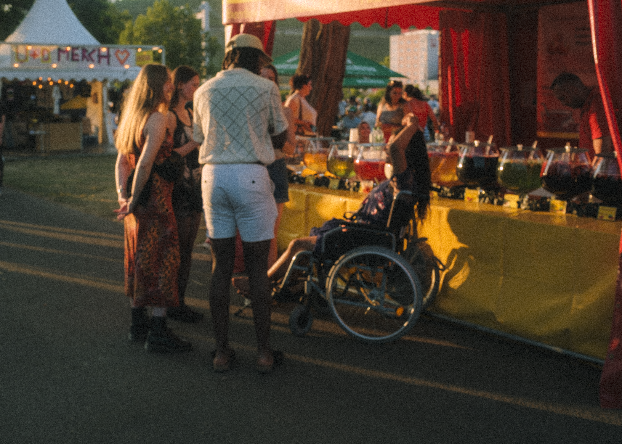 A group of young women and a person in a wheelchair gather around a brightly colored fairground game stall at a festival during the evening. The stall features large glass containers filled with various colored liquids, possibly for a game or drink. 