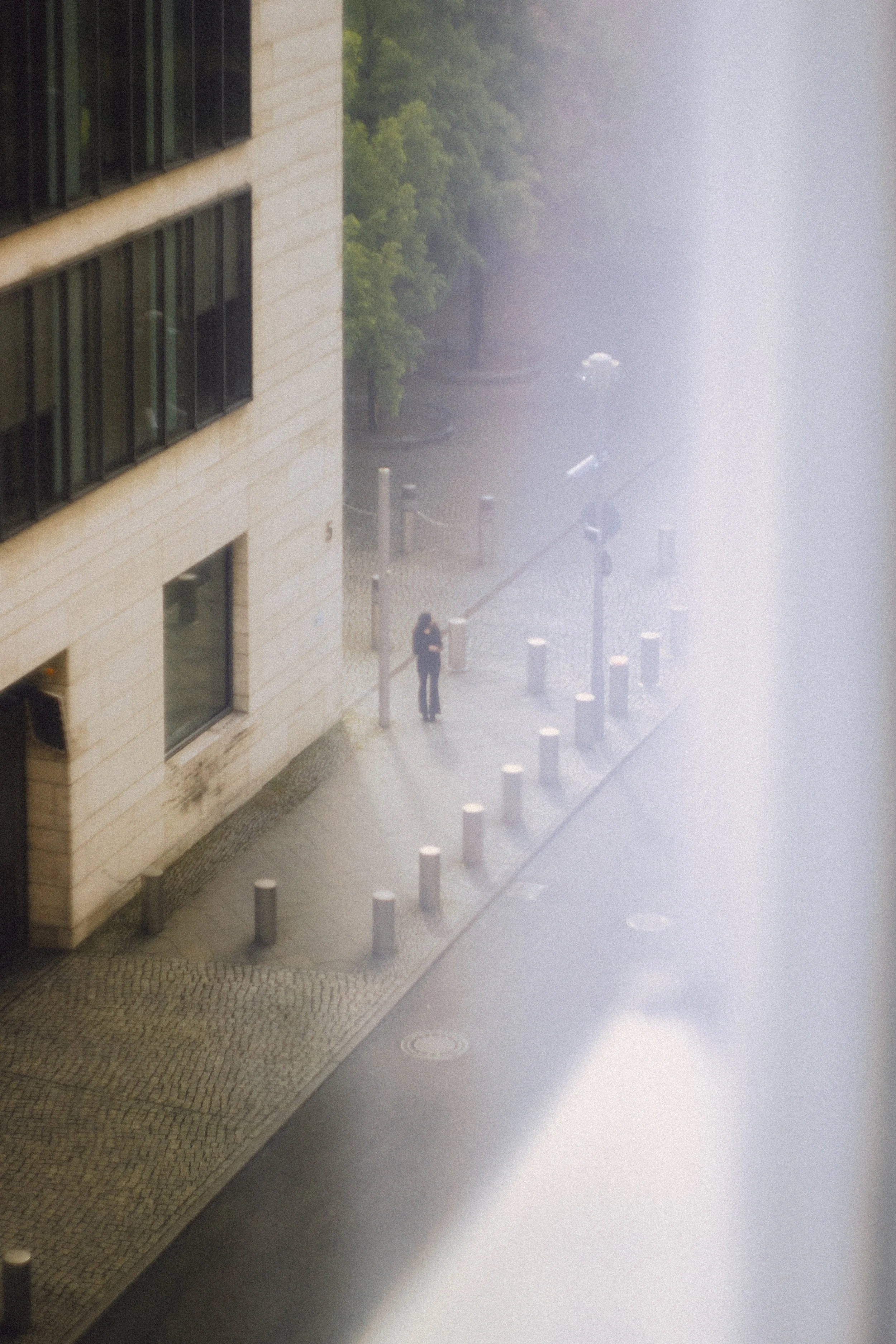 A person standing on a city sidewalk near a building, captured through a window with water or condensation blurring part of the view.