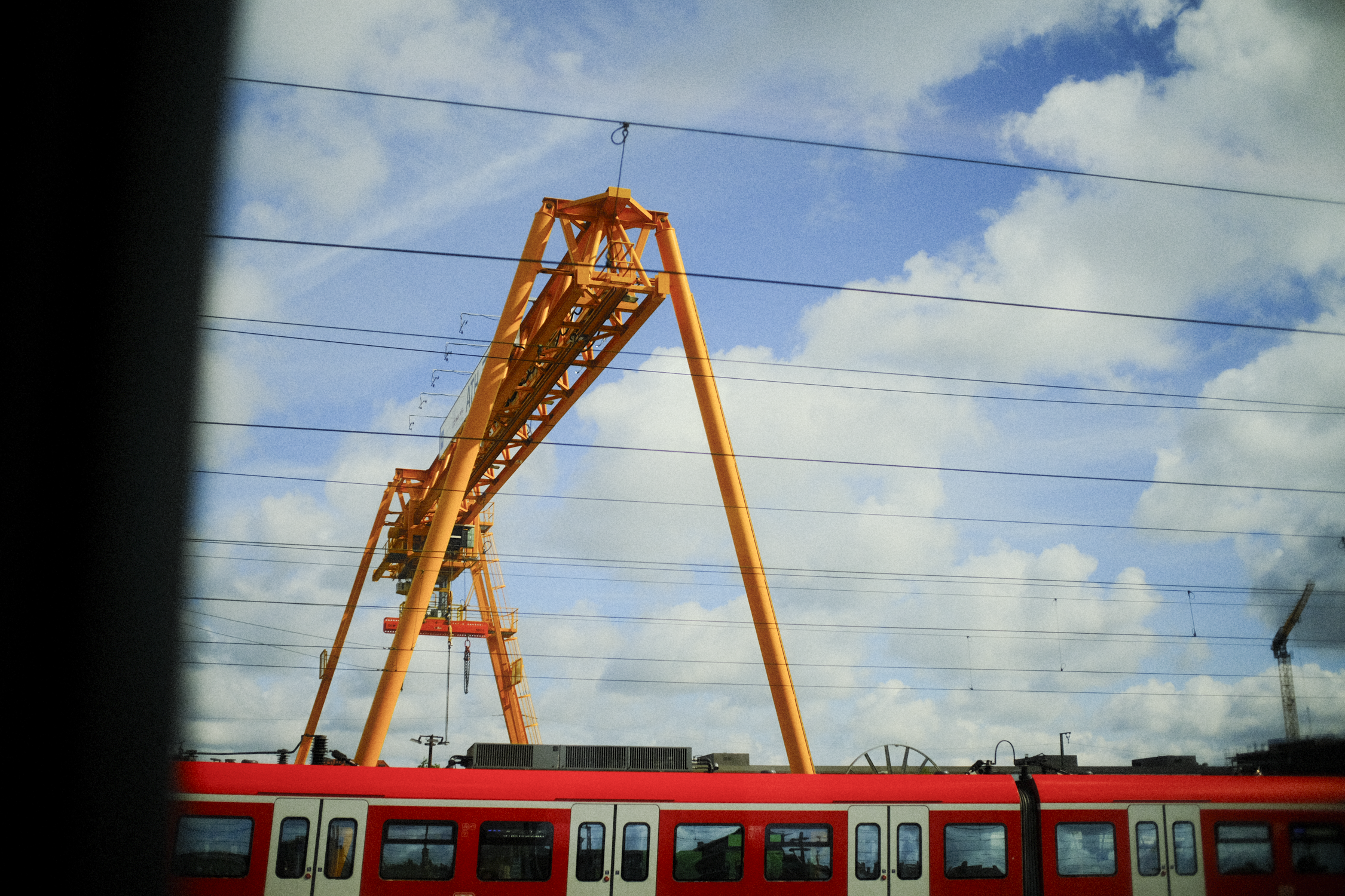 An orange construction crane behind a red train with a blue sky and white clouds.
