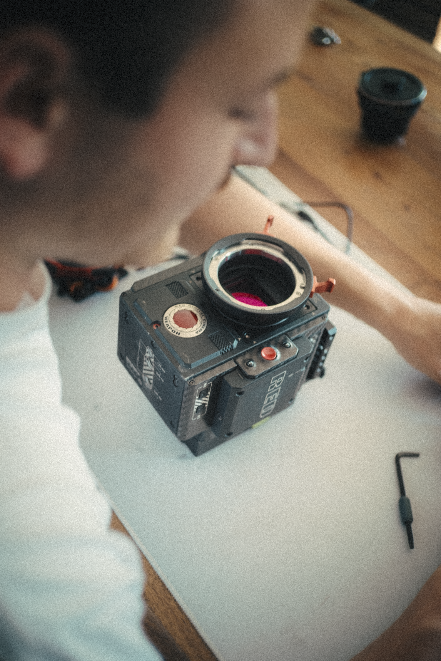 A person working with a large camera on a white desk, with a small black container and a small tool nearby.